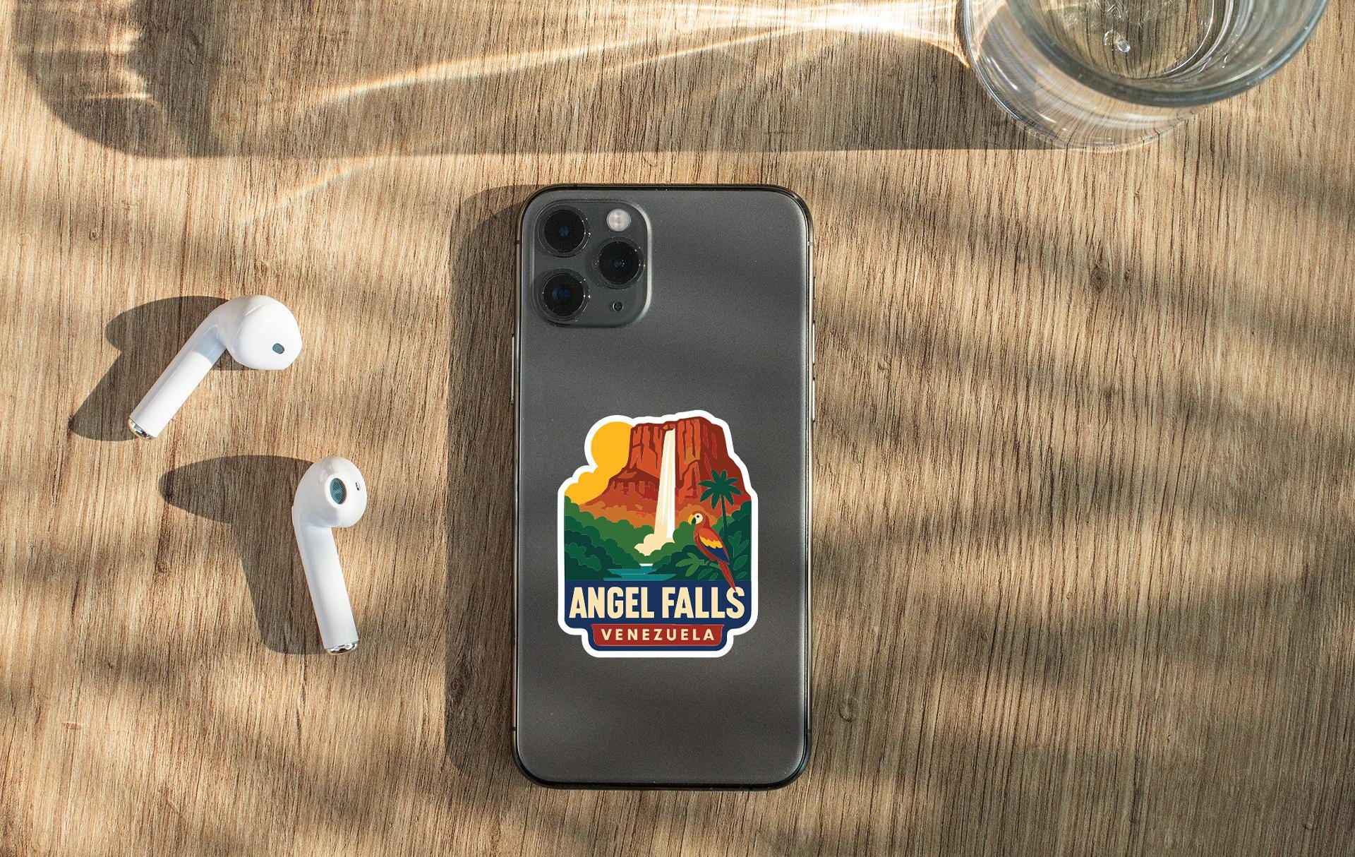 a smartphone with a sticker of Angel Falls, a famous waterfall located in Venezuela, placed on a wooden surface alongside a pair of white earbuds and a glass of water.
