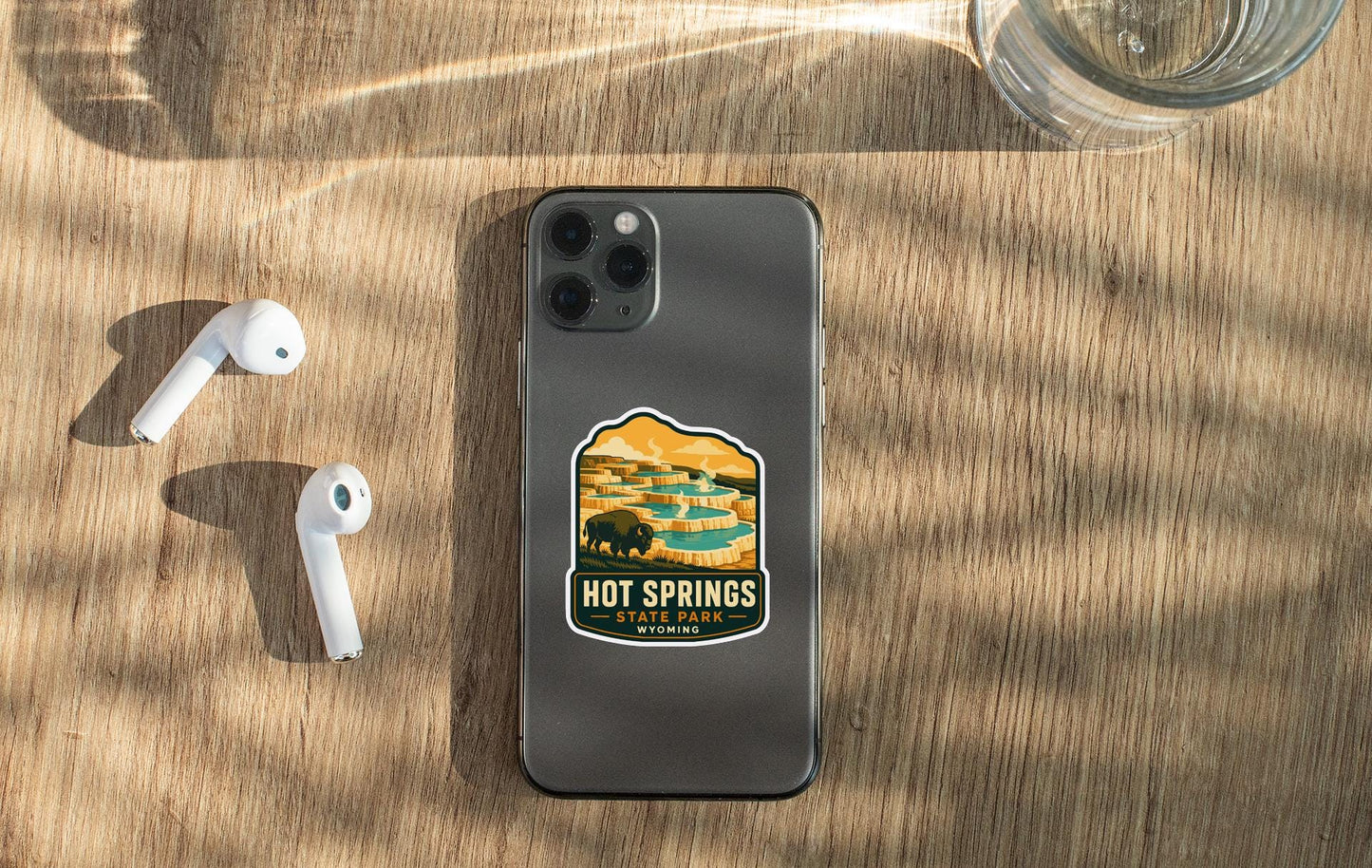a smartphone with a sticker on it, placed on a wooden surface alongside a pair of white earbuds and a glass of water.