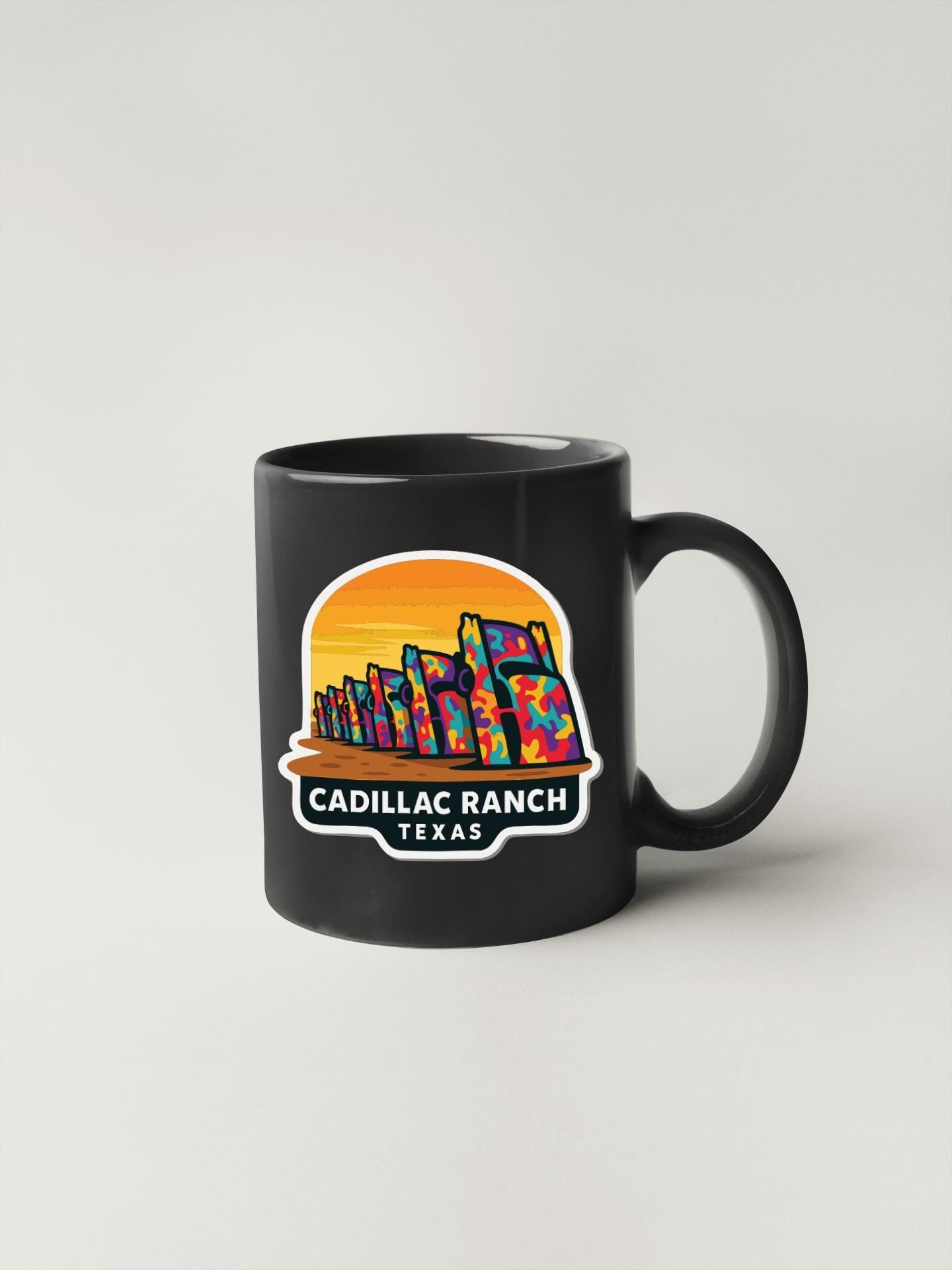 A black coffee mug with a colorful graphic design depicting a sunset over a city skyline, with the text "CADILLAC RANCH TEXAS" printed on it.