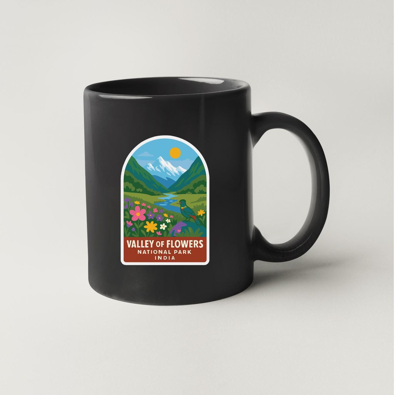 A black coffee mug with a colorful graphic depicting a valley of flowers and mountains, along with the text "Valley of Flowers National Park, Idaho".