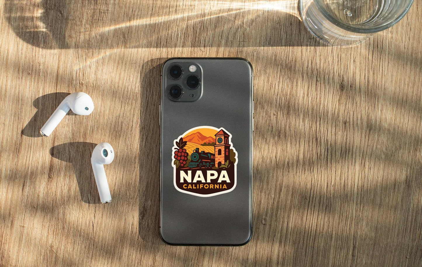 a smartphone with a sticker of Napa, California, placed on a wooden surface alongside a pair of white earbuds and a glass of water.