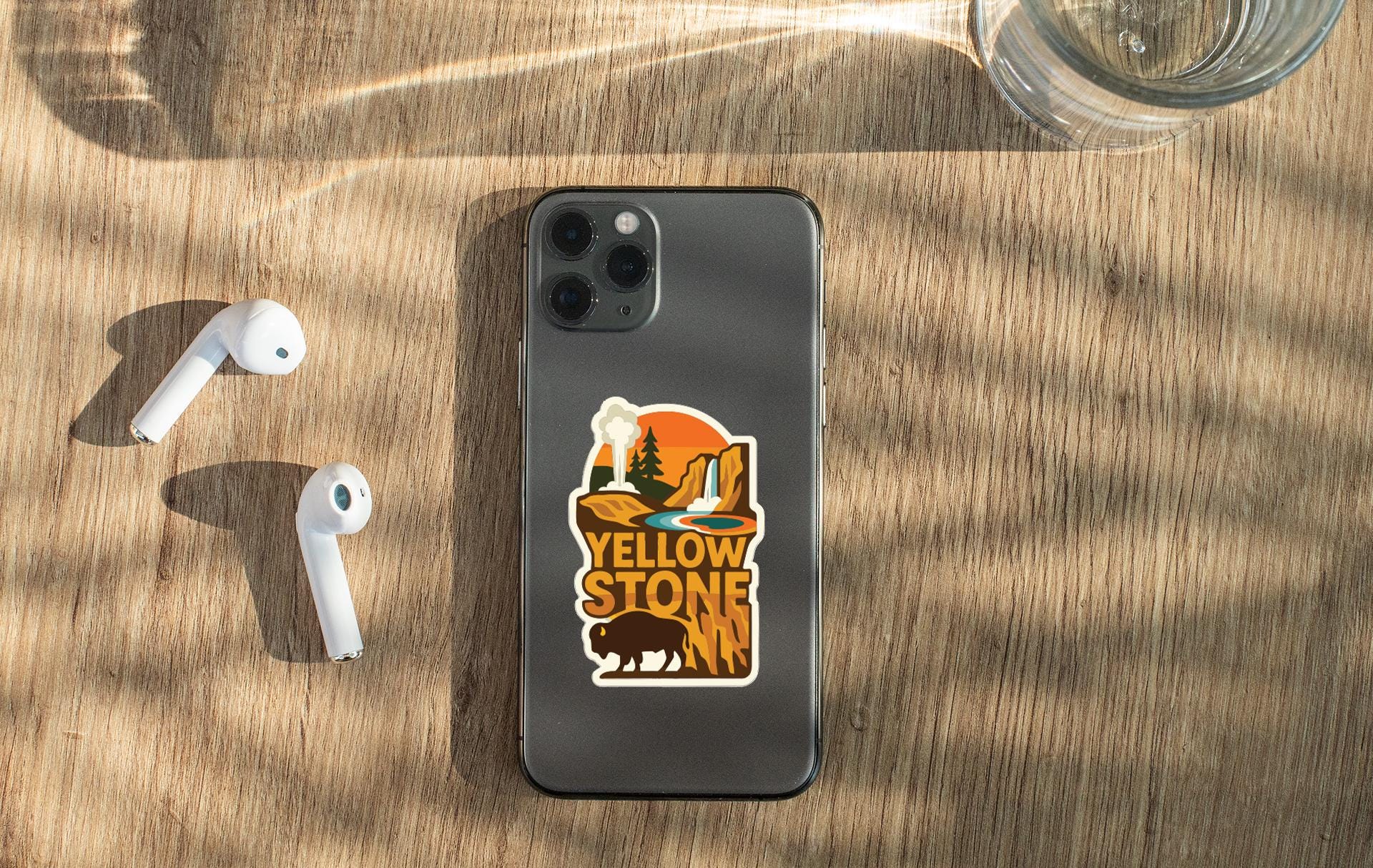 a smartphone with a sticker on the back, along with a pair of white earbuds and a glass of water on a wooden surface.