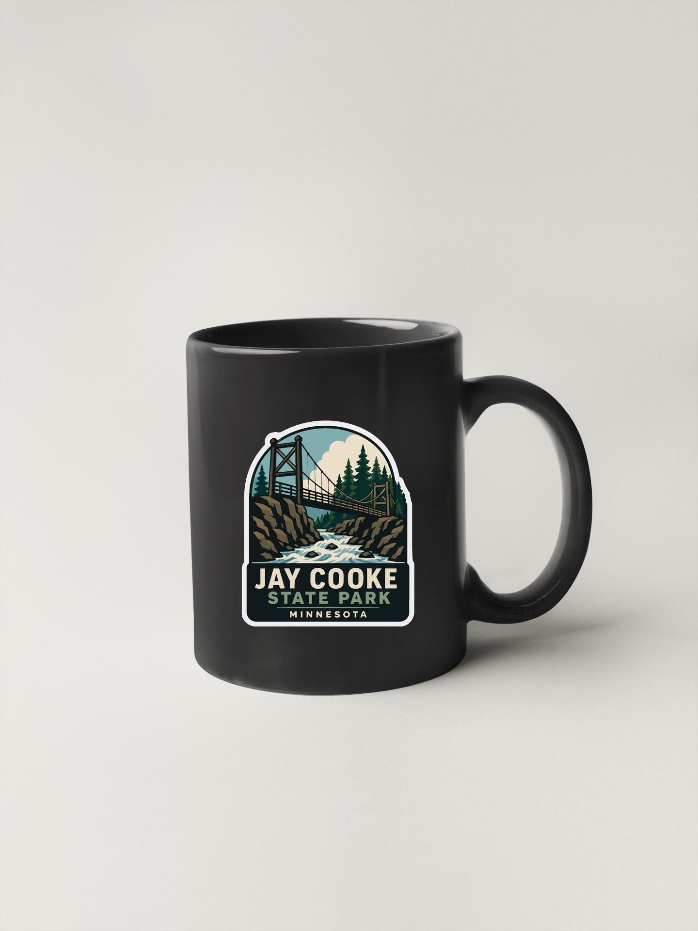 A black coffee mug with a graphic design depicting a bridge and a scenic landscape, along with the text "JAY COOOK STATE PARK MINNESOTA".