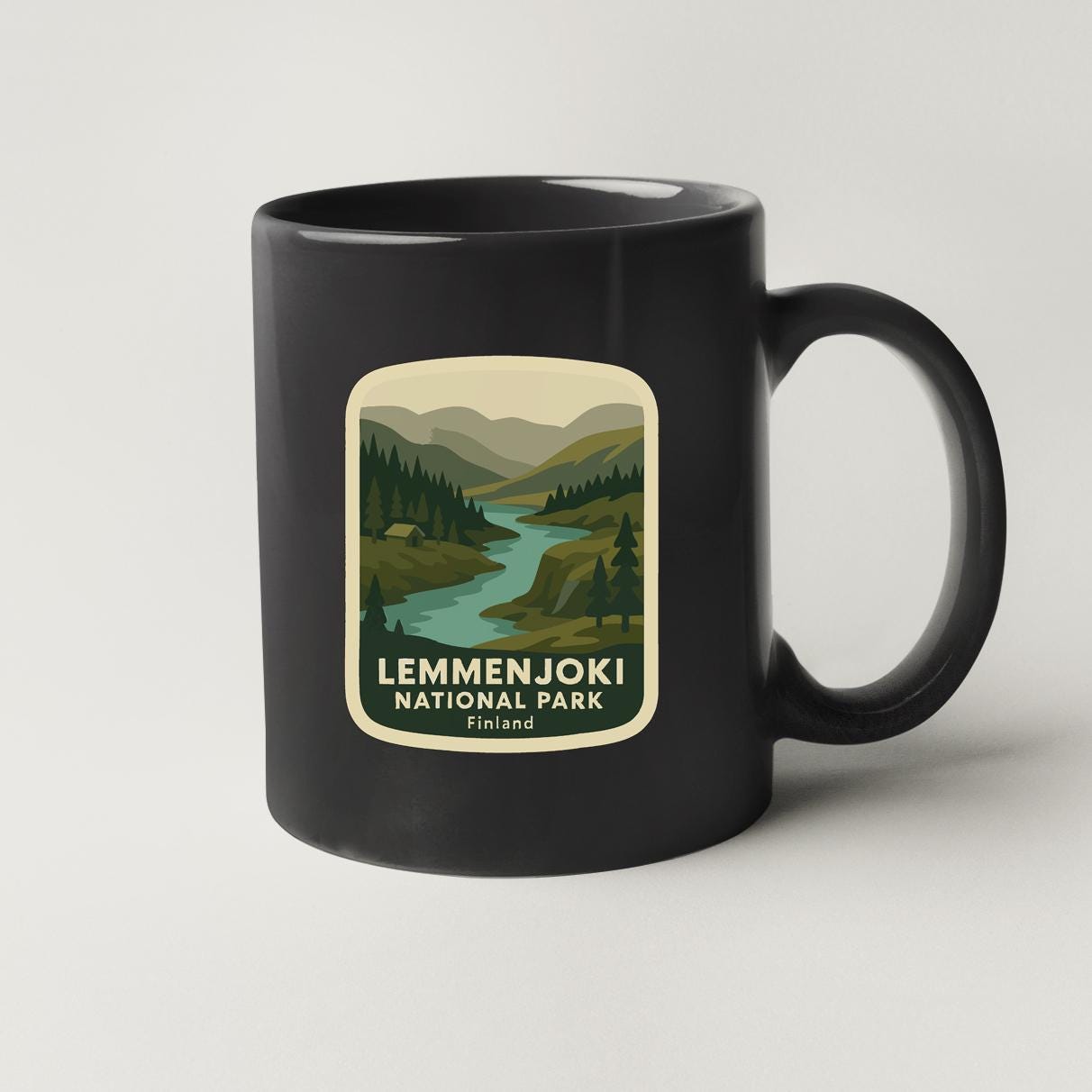 A black coffee mug with a label that says "LEMMENJOKI NATIONAL PARK" and features a scenic landscape with a river and mountains.