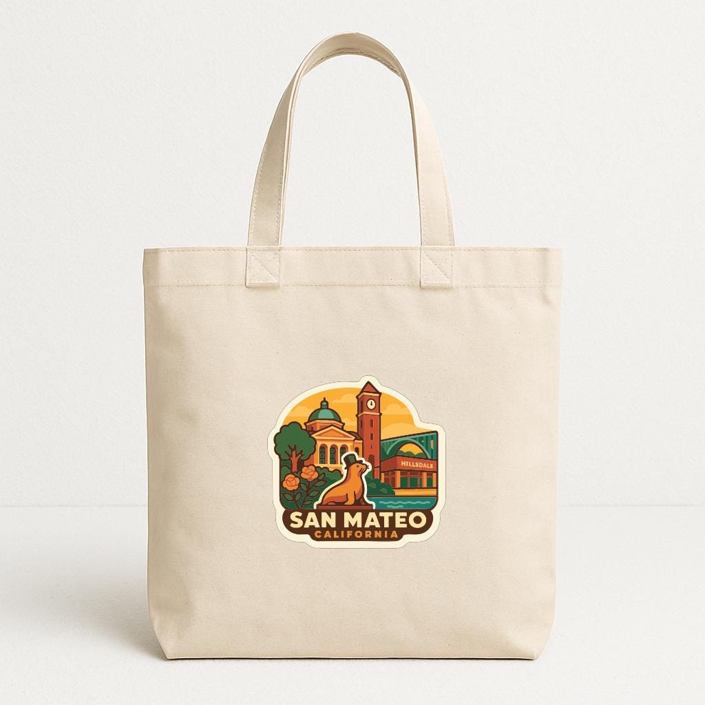 A beige tote bag with a printed design depicting a cityscape and landmarks, including a clock tower, a church, and a building, along with the text "SAN MATEO, CALIFORNIA".