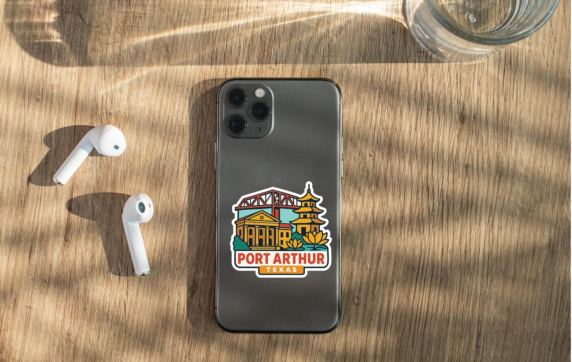 a smartphone with a sticker on it, placed on a wooden surface alongside a pair of white earbuds and a glass of water.