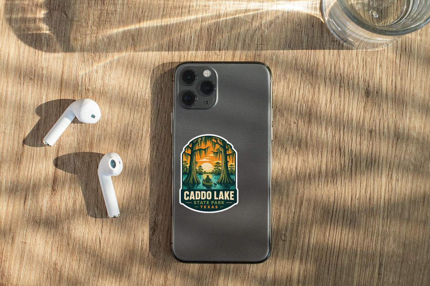 a smartphone with a sticker on it, placed on a wooden surface alongside a pair of white earbuds and a glass of water.