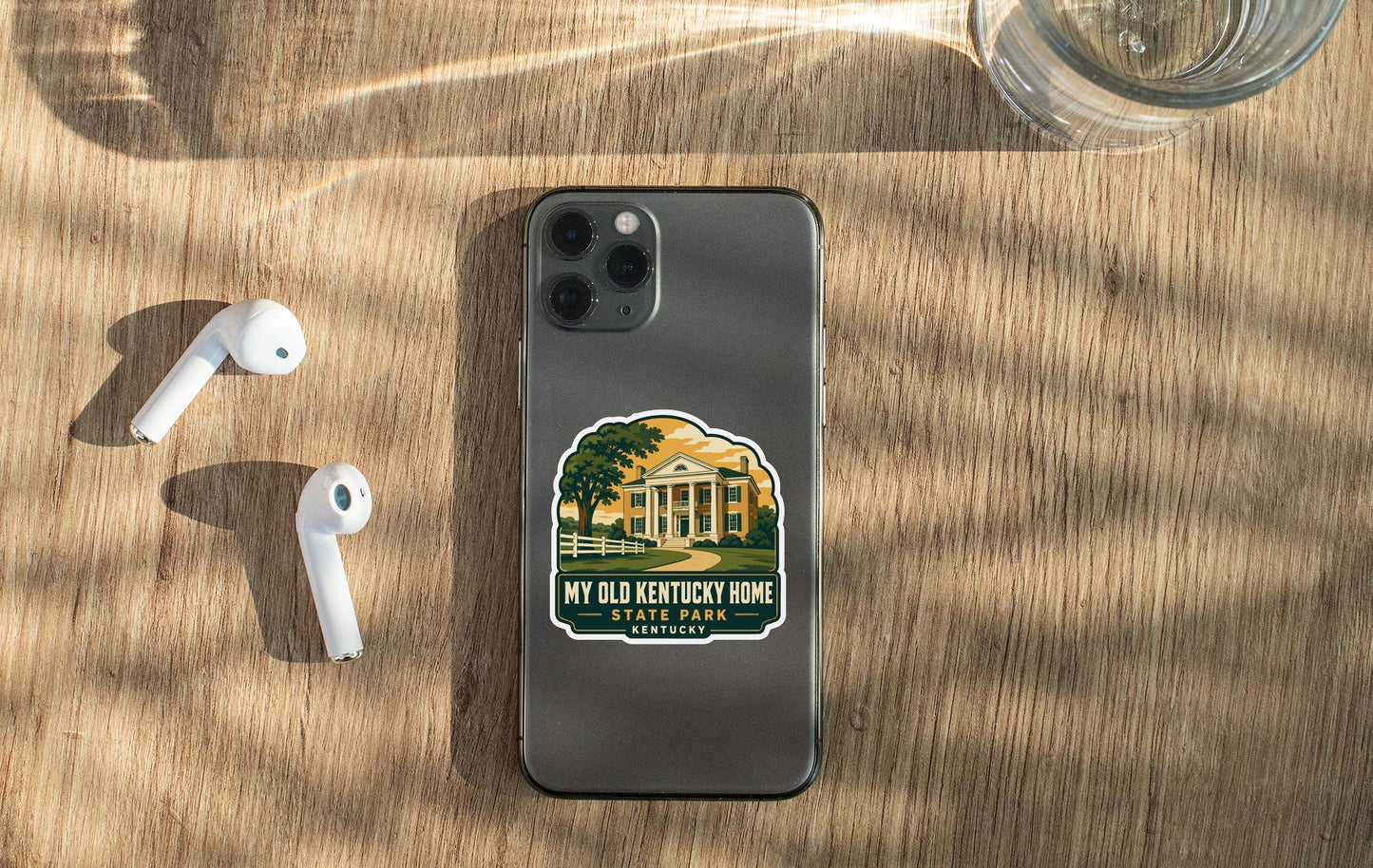 a smartphone with a sticker on the back, placed on a wooden surface alongside a pair of white earbuds and a glass of water.