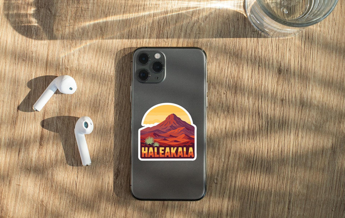 a smartphone with a sticker of a mountain and the word "Haleakala" on it, along with a pair of white earbuds and a glass of water on a wooden surface.