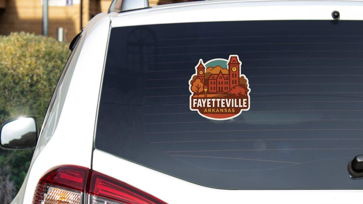 A sticker on the rear window of a white vehicle, displaying the text "FAYETTEVILLE ARKANSAS" along with an illustration of a building and a clock tower.