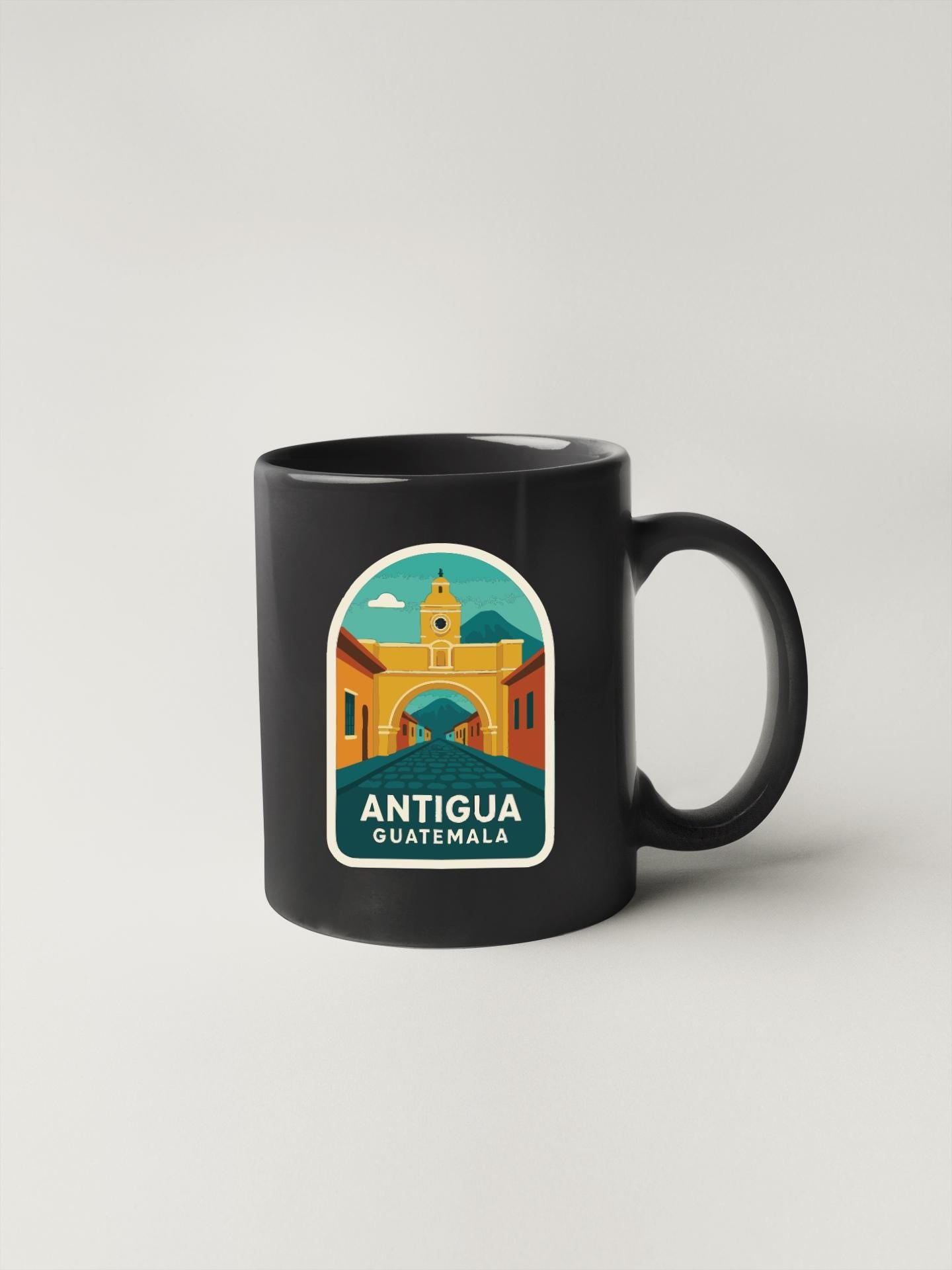 A black coffee mug with a colorful illustration of Antigua, Guatemala, featuring a yellow building with a clock tower and a blue sky with clouds.