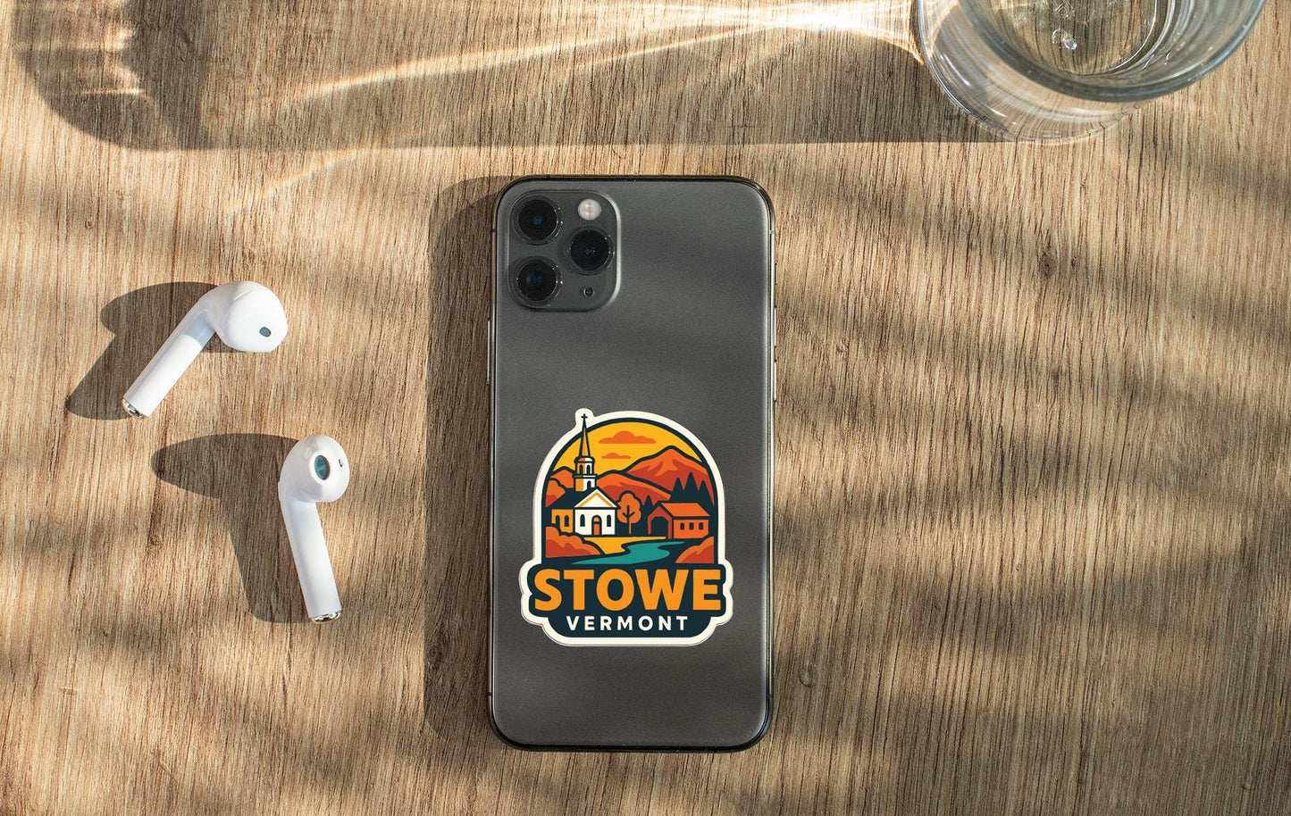 a smartphone with a Stow Vermont sticker on the back cover, placed on a wooden surface alongside a pair of white earbuds and a glass of water.