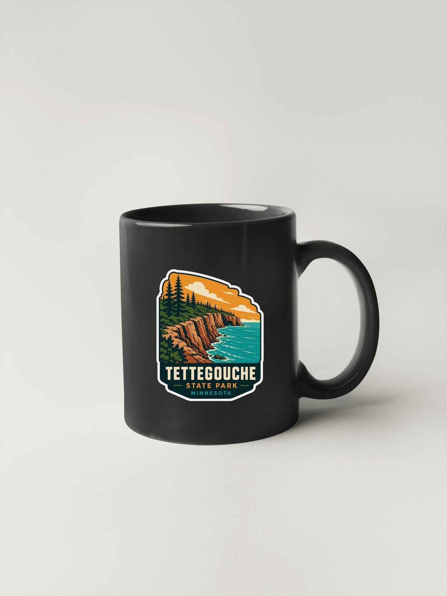 A black coffee mug with a graphic design depicting a scenic landscape, including a cliff overlooking a body of water and trees in the background.