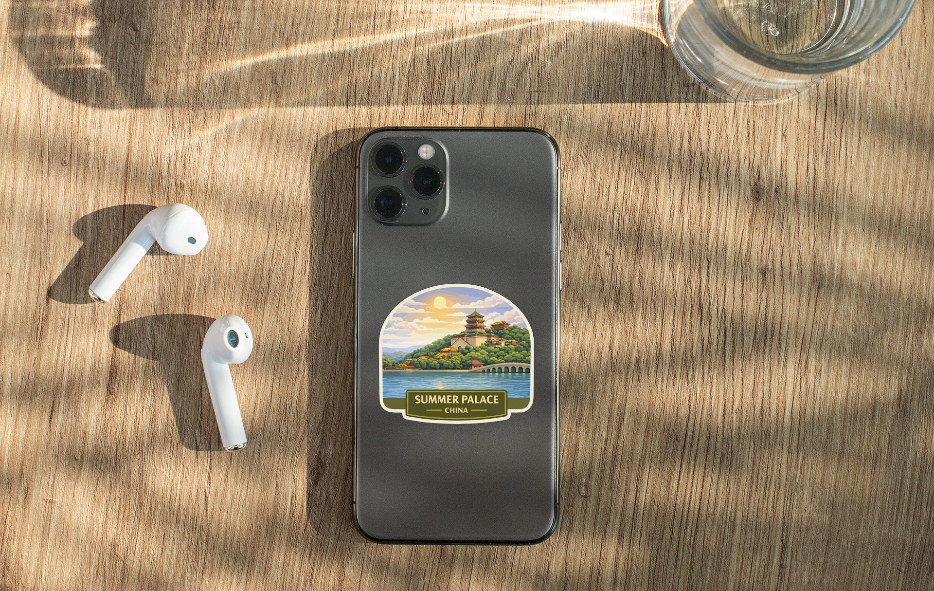 a smartphone with a sticker of a scenic view, a pair of white earbuds, and a glass of water on a wooden surface.