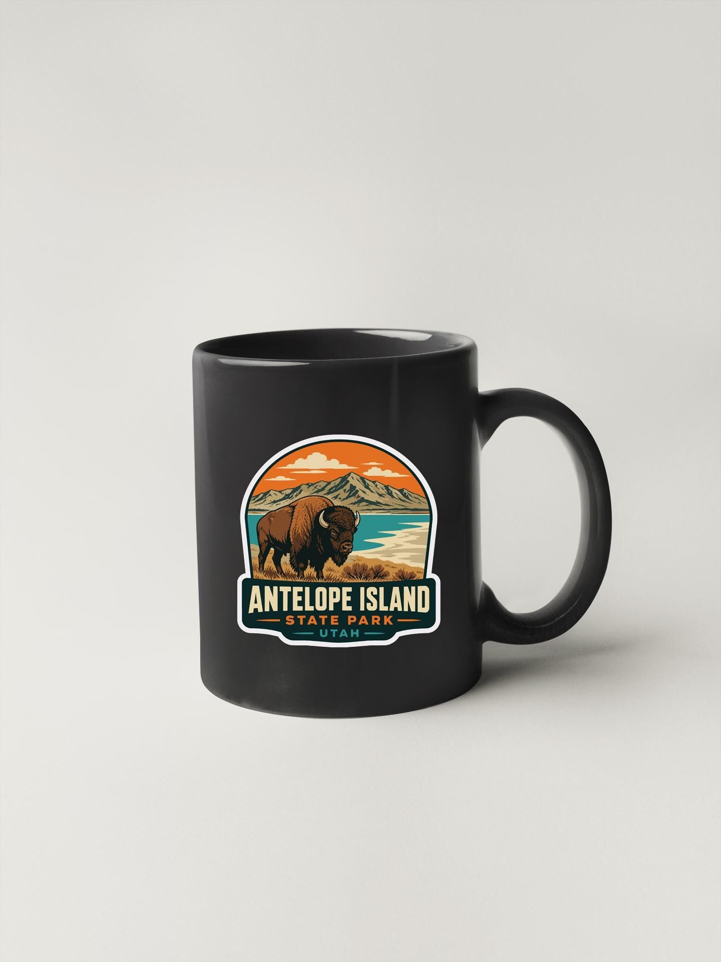 A black coffee mug with a graphic design depicting a bison in a scenic landscape, along with the text "ANTELOPE ISLAND STATE PARK".