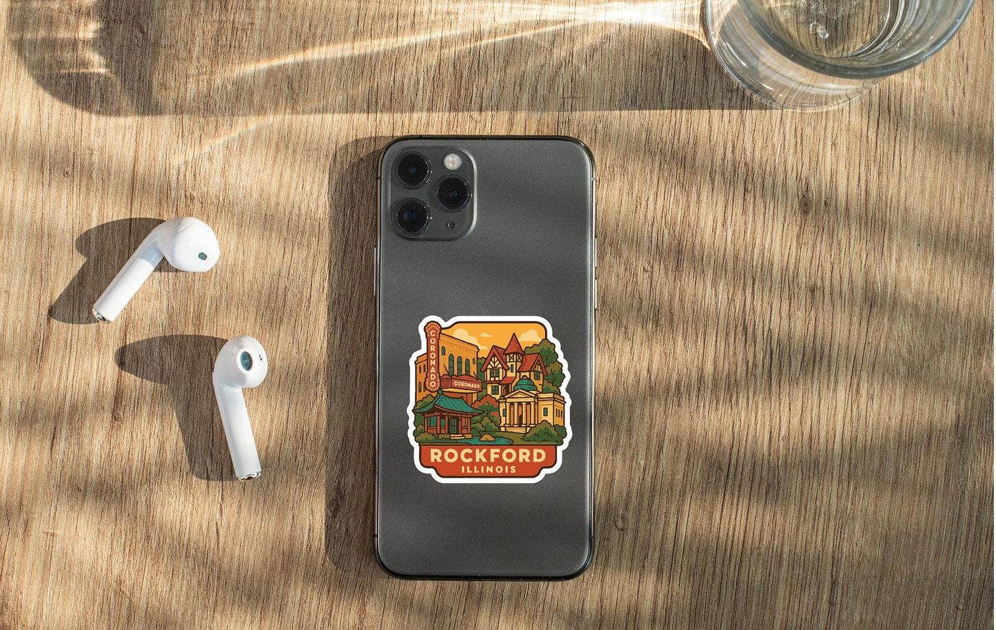 a smartphone with a sticker of Rockford, Illinois, on its back, along with a pair of white earbuds and a glass of water on a wooden surface.