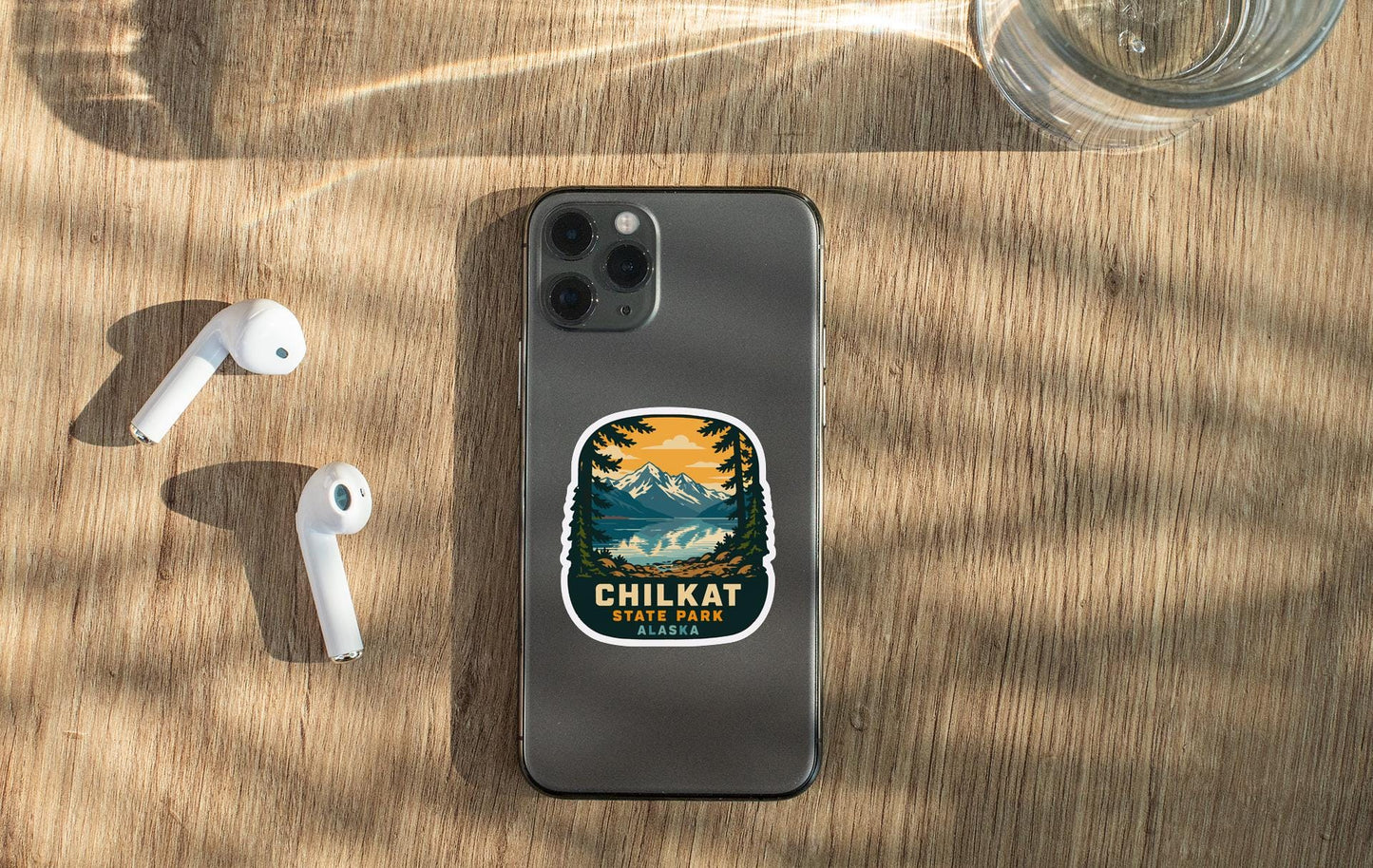 a smartphone with a sticker on it, placed on a wooden surface alongside a pair of white earbuds and a glass of water.