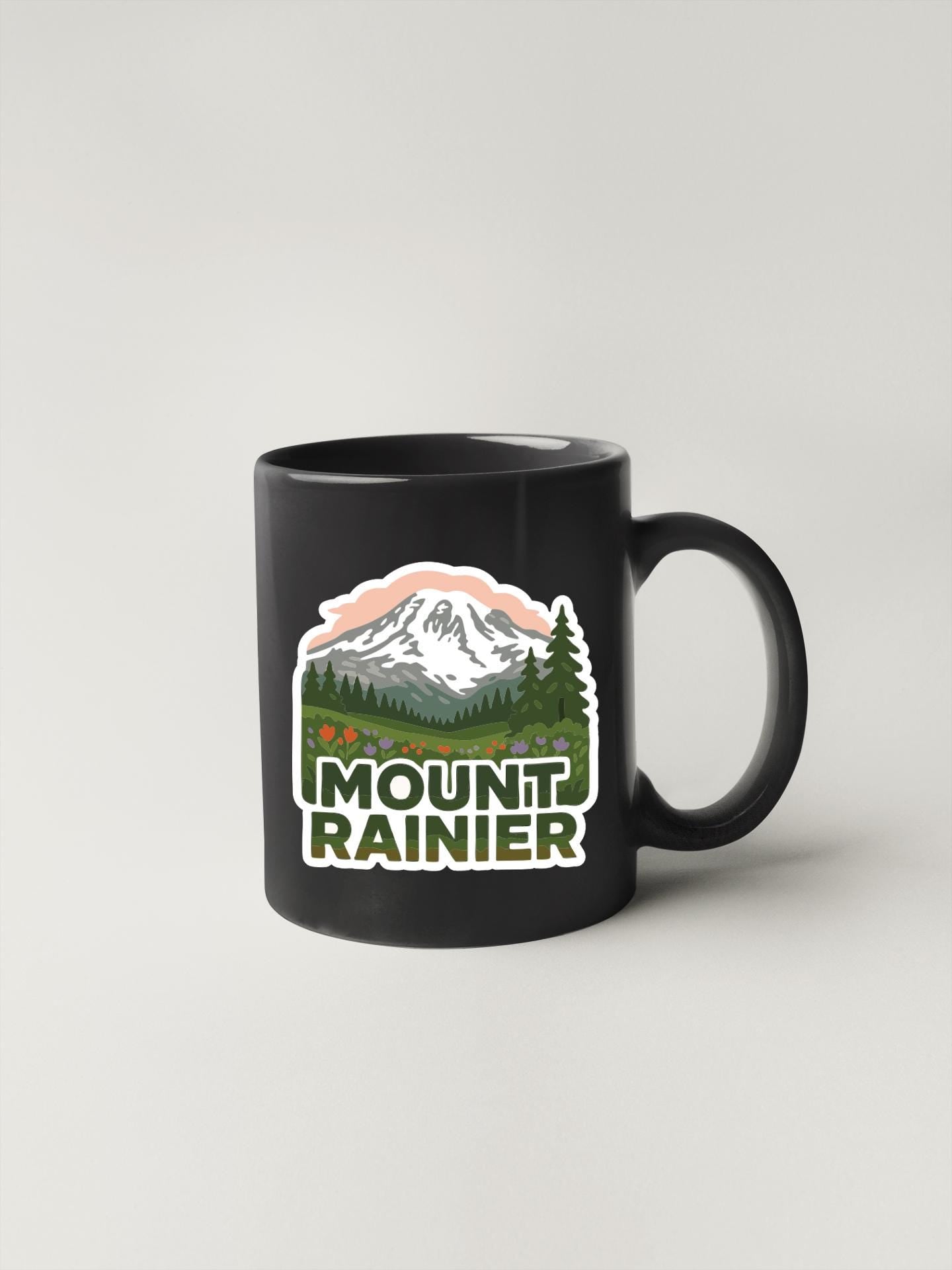 A black coffee mug with a graphic design featuring a mountain landscape and the text "MOUNT RAINIER" printed on it.