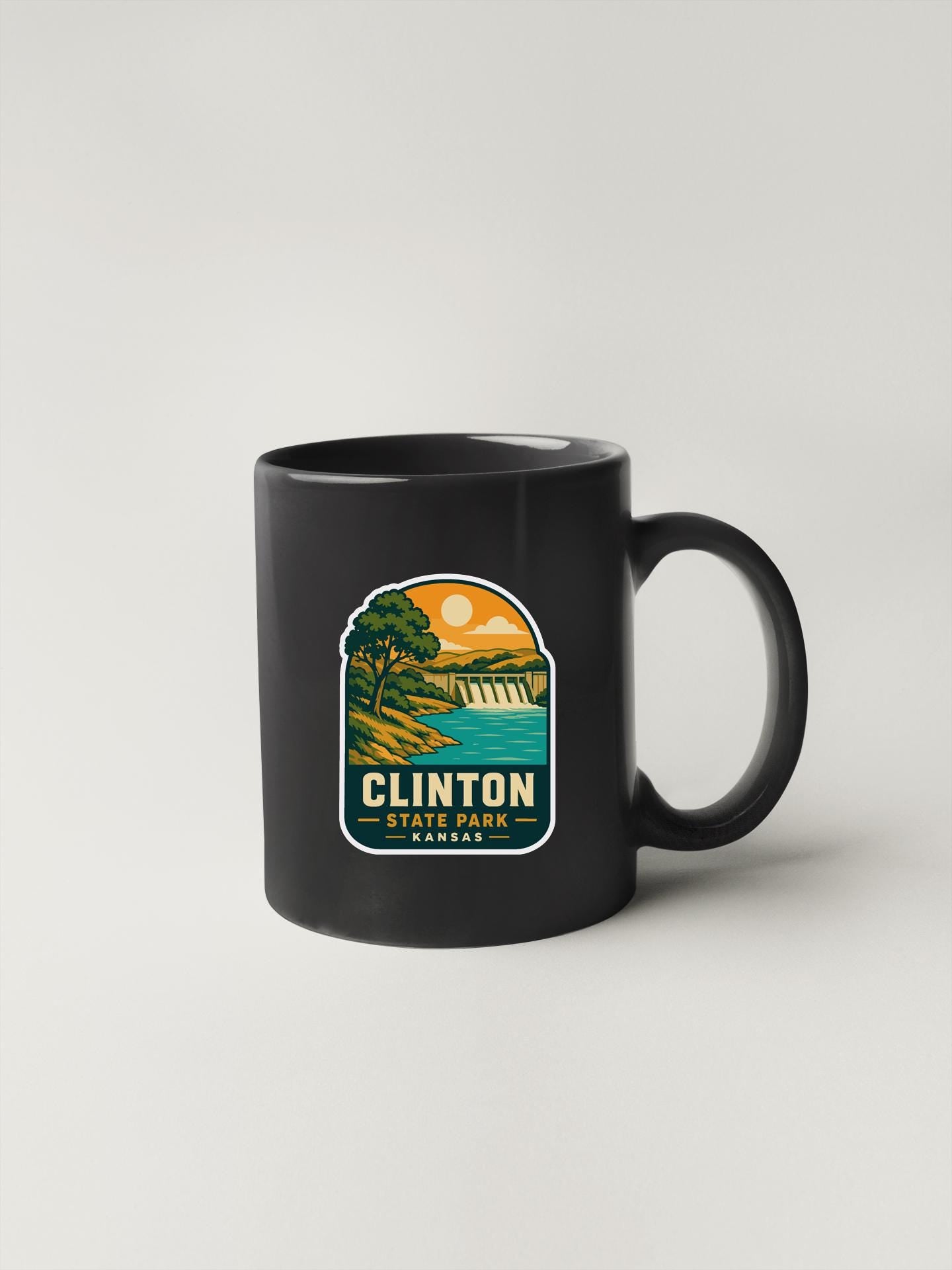 A black coffee mug with a graphic design depicting a scenic landscape, including a lake, mountains, and a sunset.