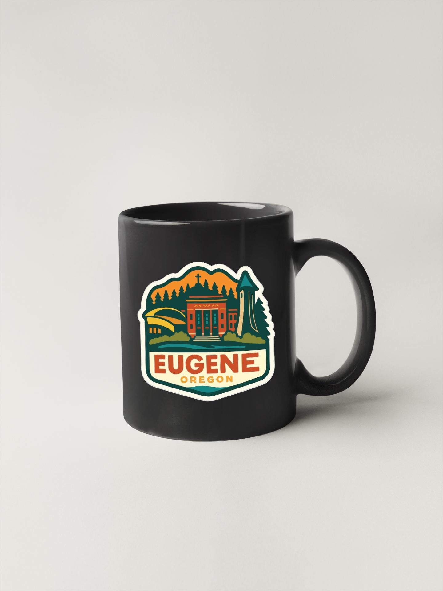 A black coffee mug with a colorful graphic depicting Eugene, Oregon, featuring a mountain, buildings, and trees.