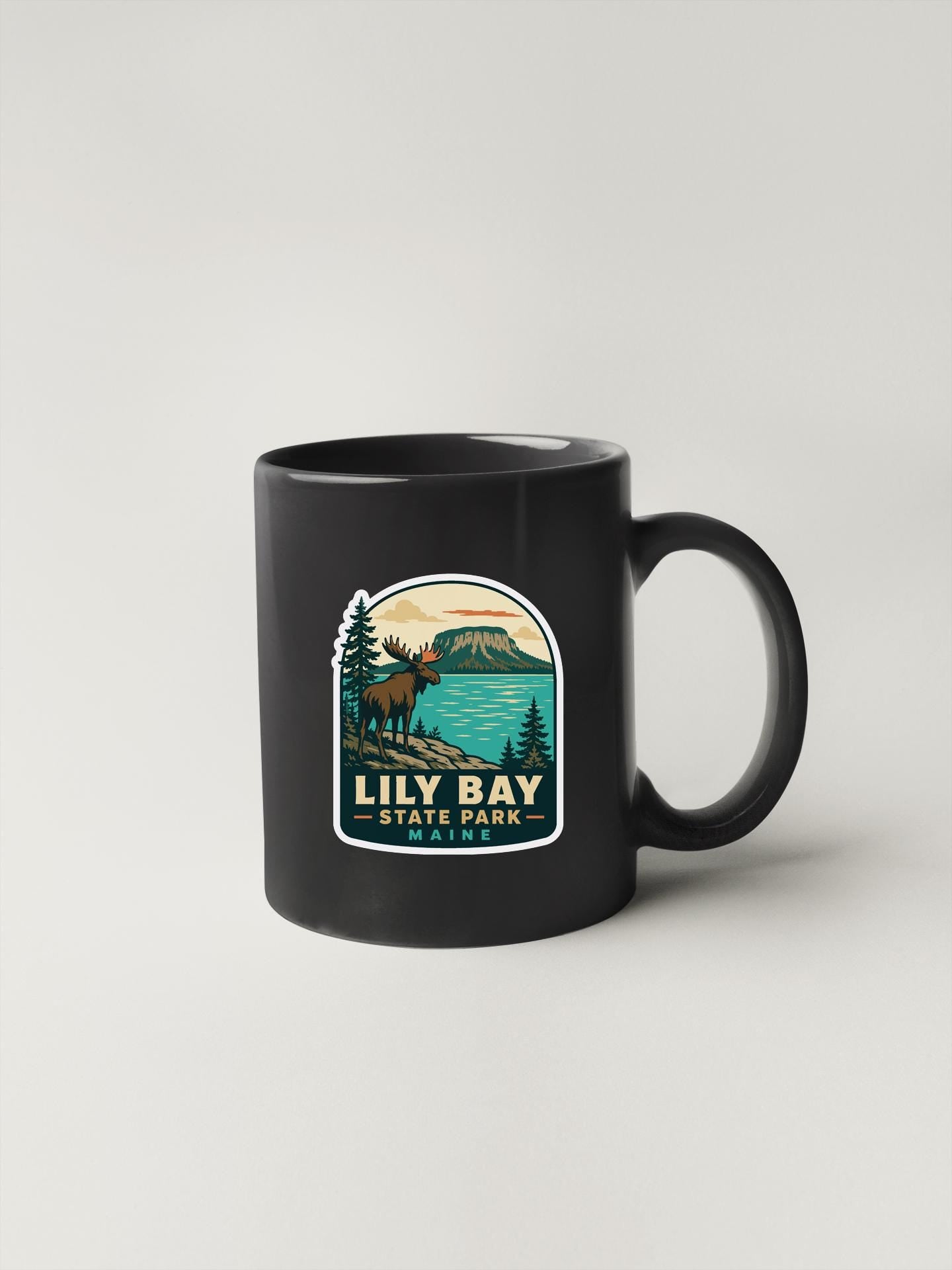 A black coffee mug with a graphic design depicting a moose and a lake, along with the text "Lily Bay State Park".