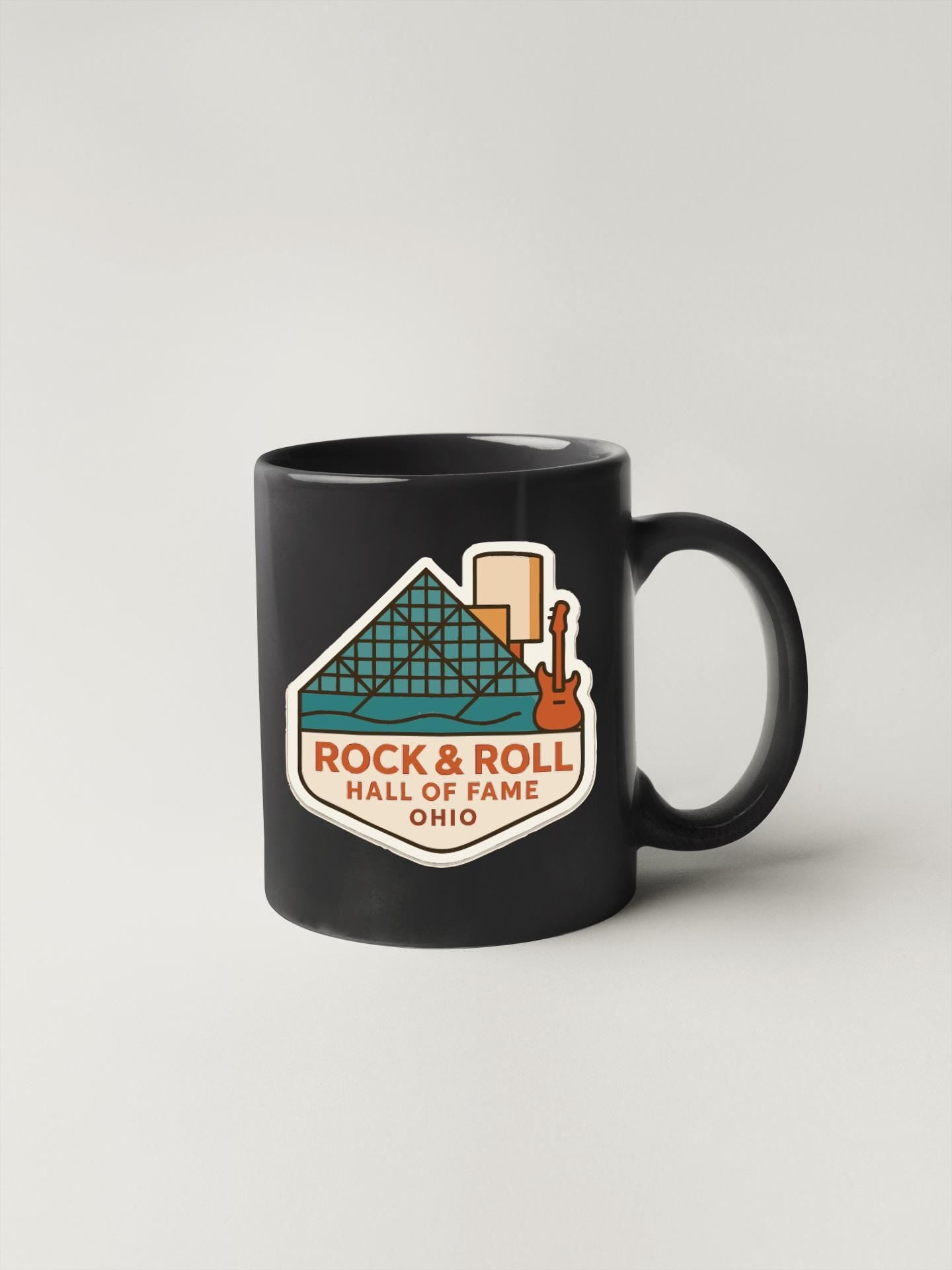 A black coffee mug with a graphic design featuring a building and a guitar, along with the text "ROCK & ROLL HALL OF FAME OHIO".