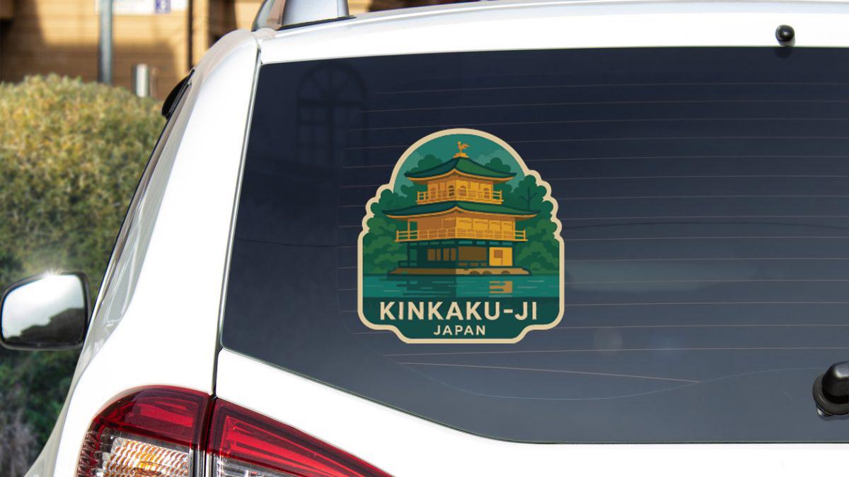A sticker on the rear window of a white car, depicting a temple with a green roof and yellow walls, along with the text "KINKAU-JI JAPAN".