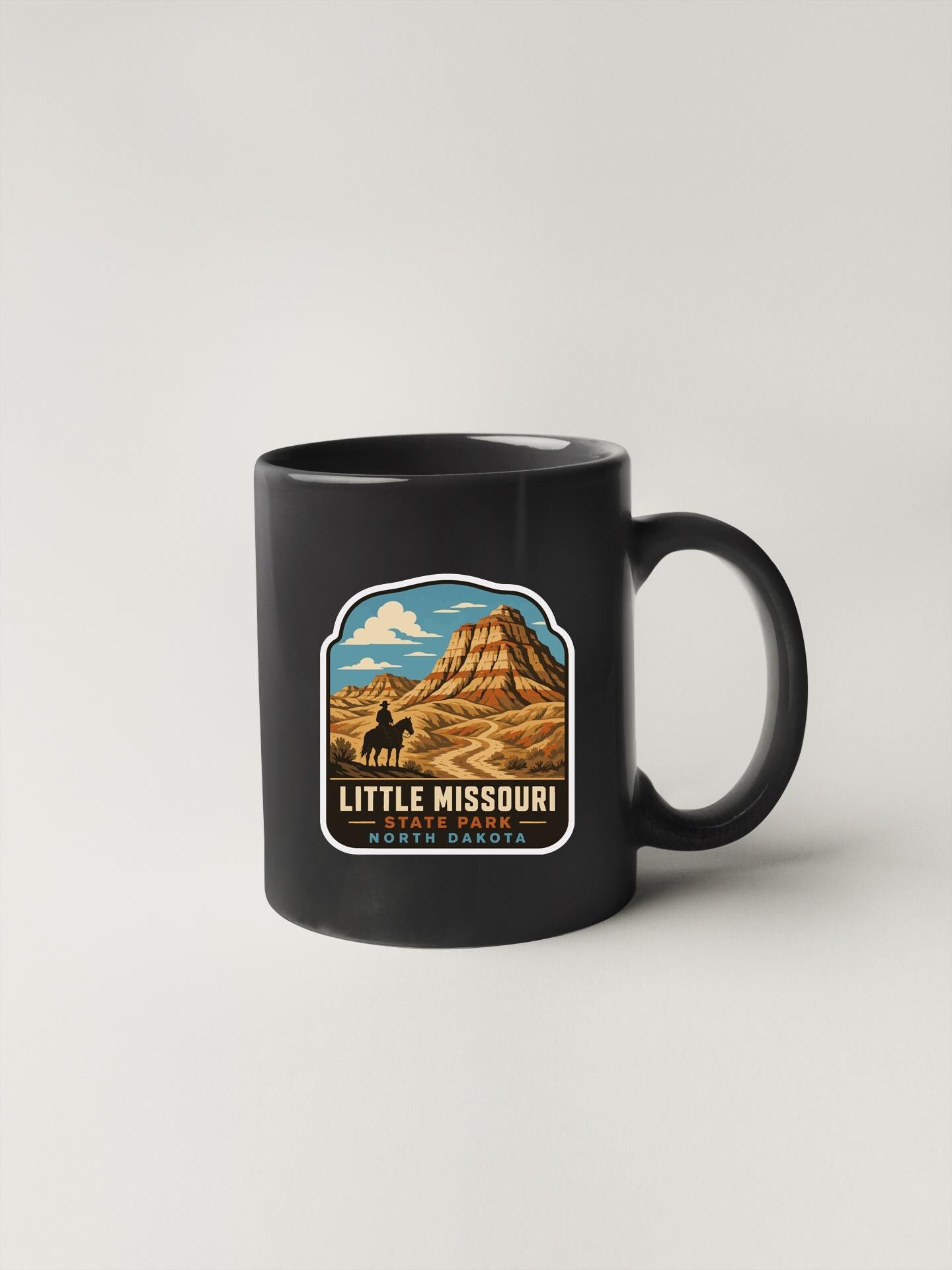 A black coffee mug with a graphic design depicting a mountain landscape and a cowboy on horseback, along with the text "LITTLE MISSOURI NORTH CAROLINA".