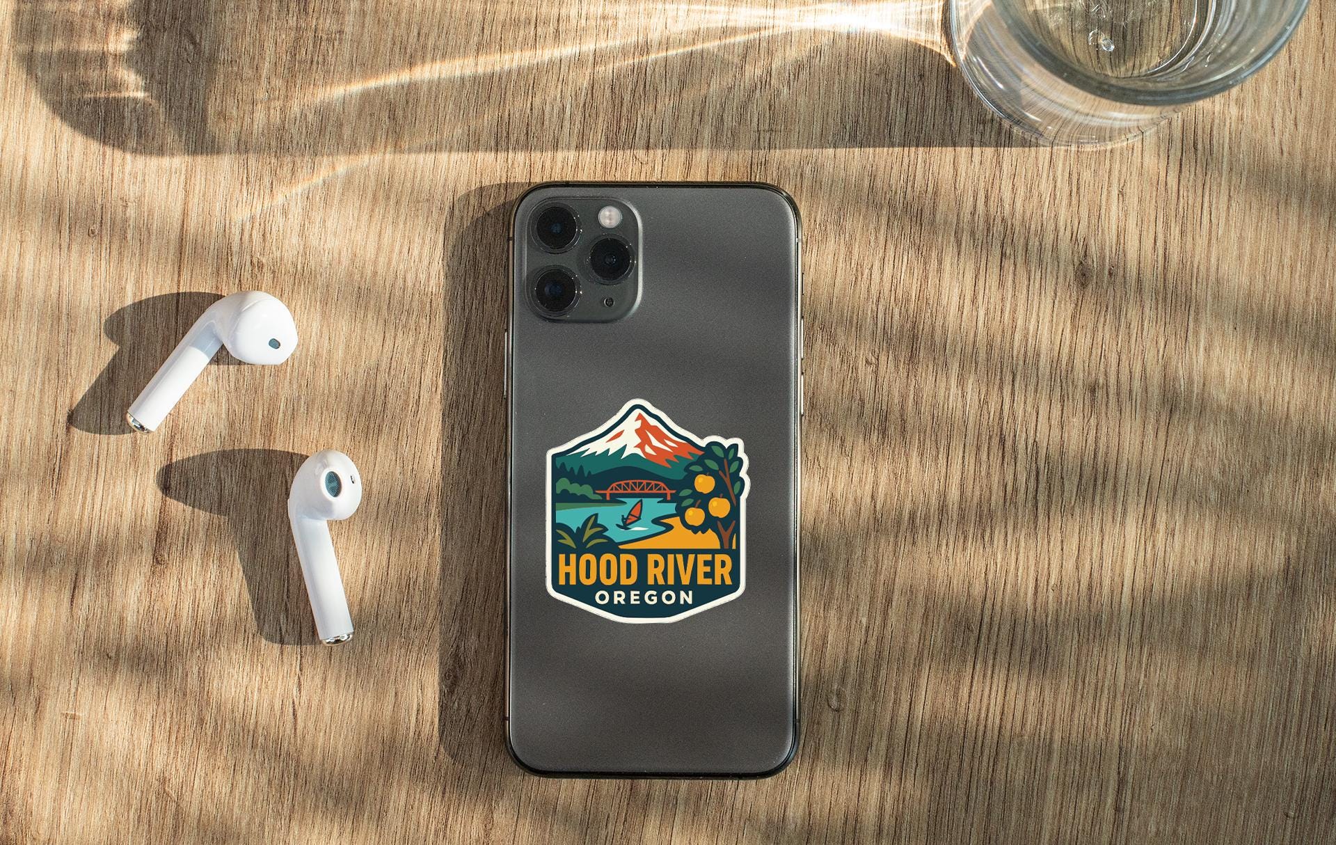a smartphone with a sticker on the back, placed on a wooden surface alongside a pair of white earbuds and a glass of water.