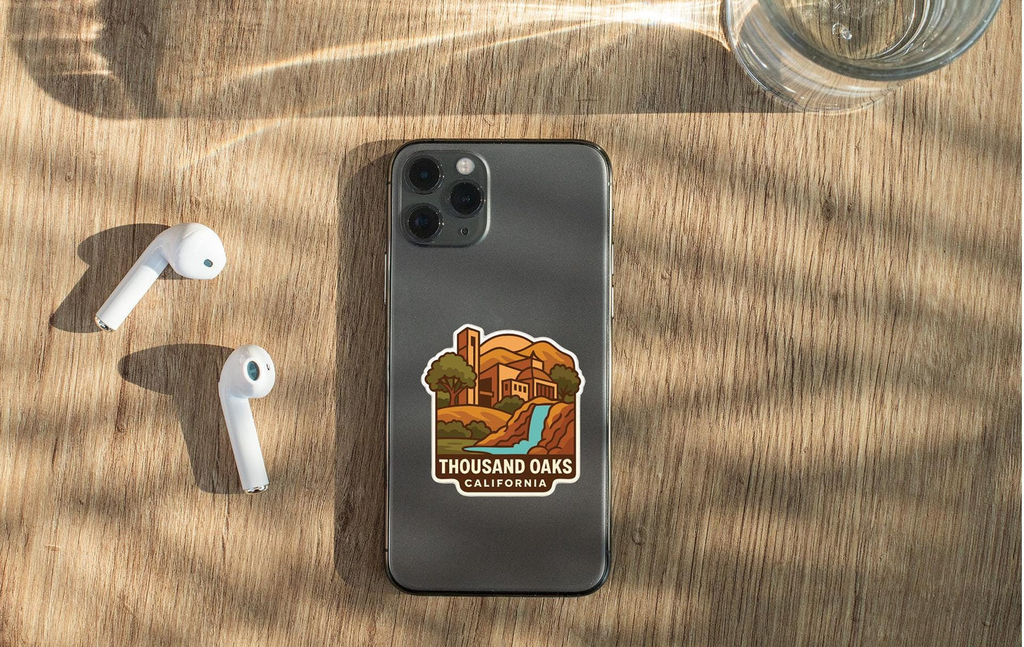 a smartphone with a sticker on the back, placed on a wooden surface alongside a pair of white earbuds and a glass of water.