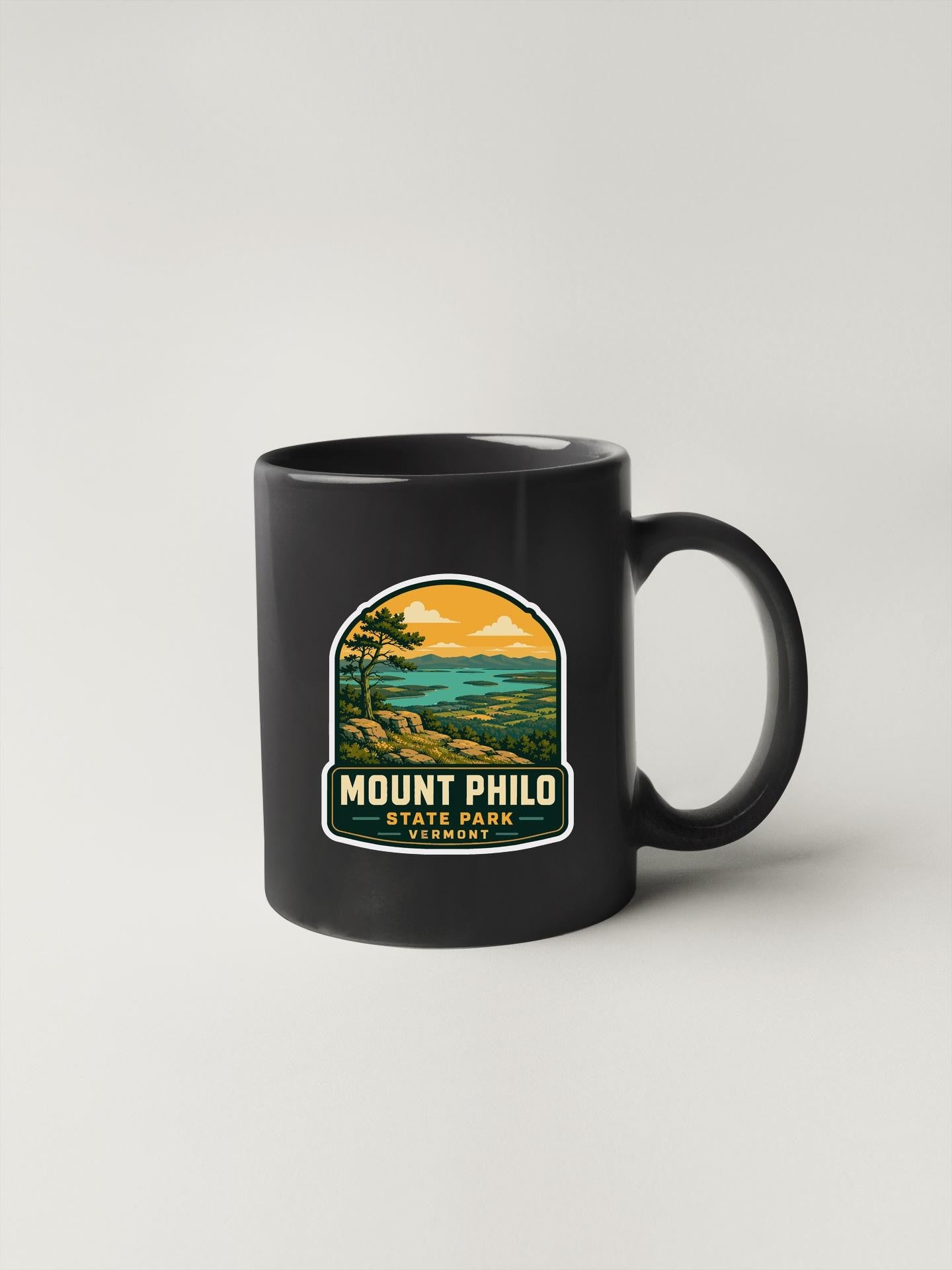 A black coffee mug with a graphic design depicting a scenic landscape, including mountains, trees, and a body of water.