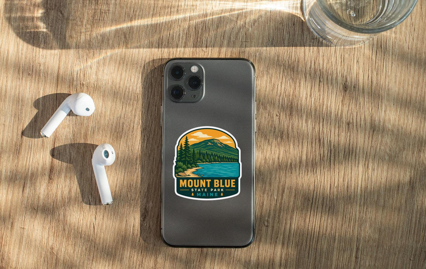 a smartphone with a sticker on the back, placed on a wooden surface alongside a pair of white earbuds and a glass of water.