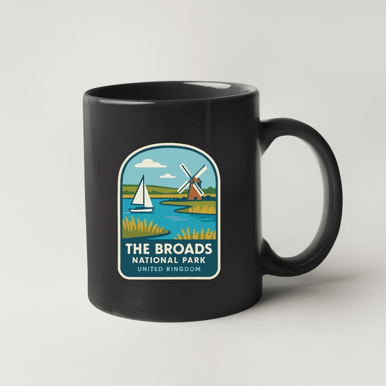 A black coffee mug with a graphic design depicting a sailboat on a lake, windmills, and a castle, along with the text "THE BROADS NATIONAL PARK UNITED KINGDOM".