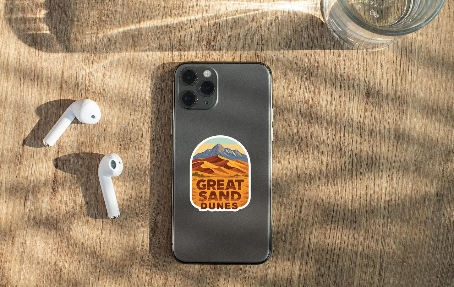 a smartphone with a sticker on the back, placed on a wooden surface alongside a pair of white earbuds and a glass of water.