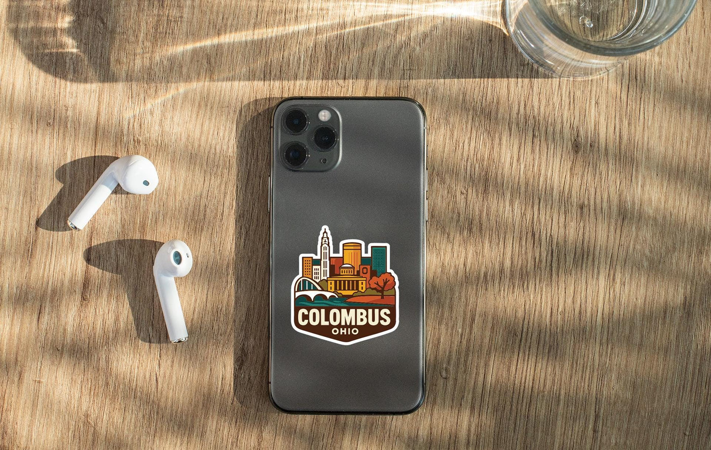 a smartphone with a sticker of the Columbus, Ohio skyline on it, along with a pair of white earbuds and a glass of water on a wooden surface.