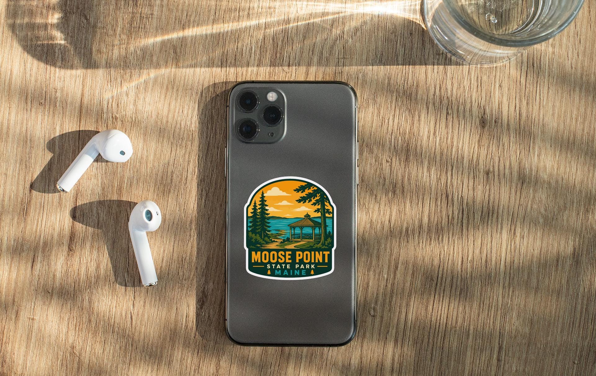 a smartphone with a sticker of Moose Point State Park on its back, along with a pair of white earbuds and a glass of water on a wooden surface.