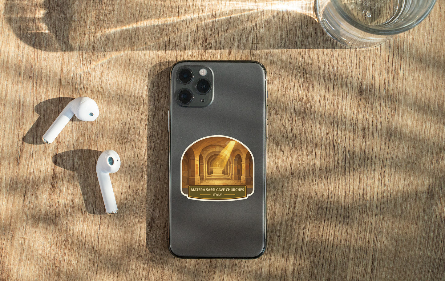 A smartphone with a sticker featuring a hallway and the text "MATERIA SARA CAESARS" is placed on a wooden surface alongside a pair of white earbuds and a glass of water.