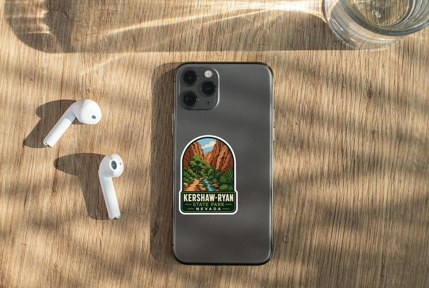 a smartphone with a sticker on the back, placed on a wooden surface alongside a pair of white earbuds and a glass of water.