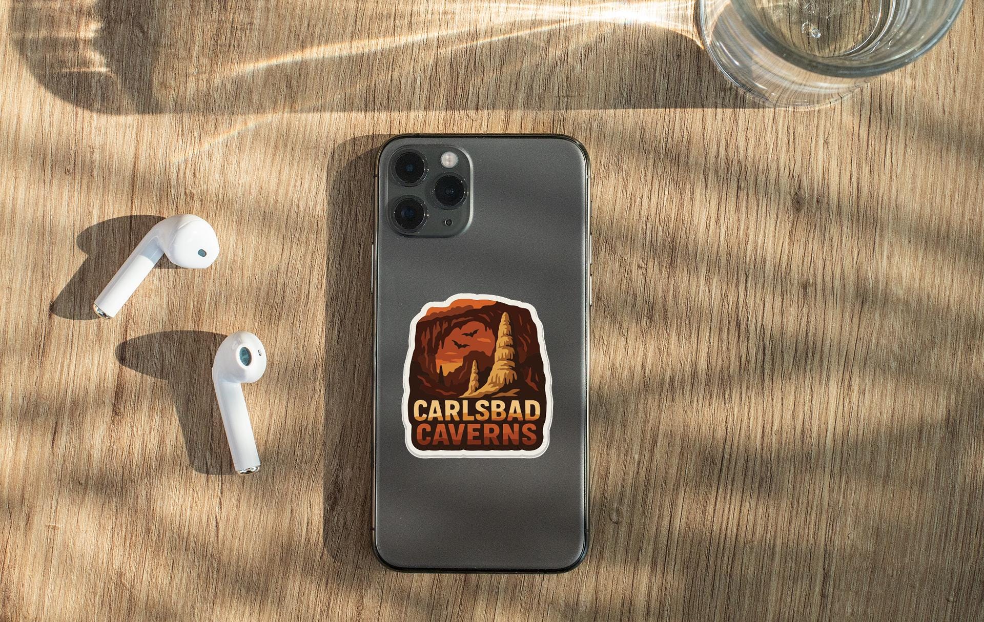 a smartphone with a sticker on it, along with a pair of white earbuds and a glass of water on a wooden surface.
