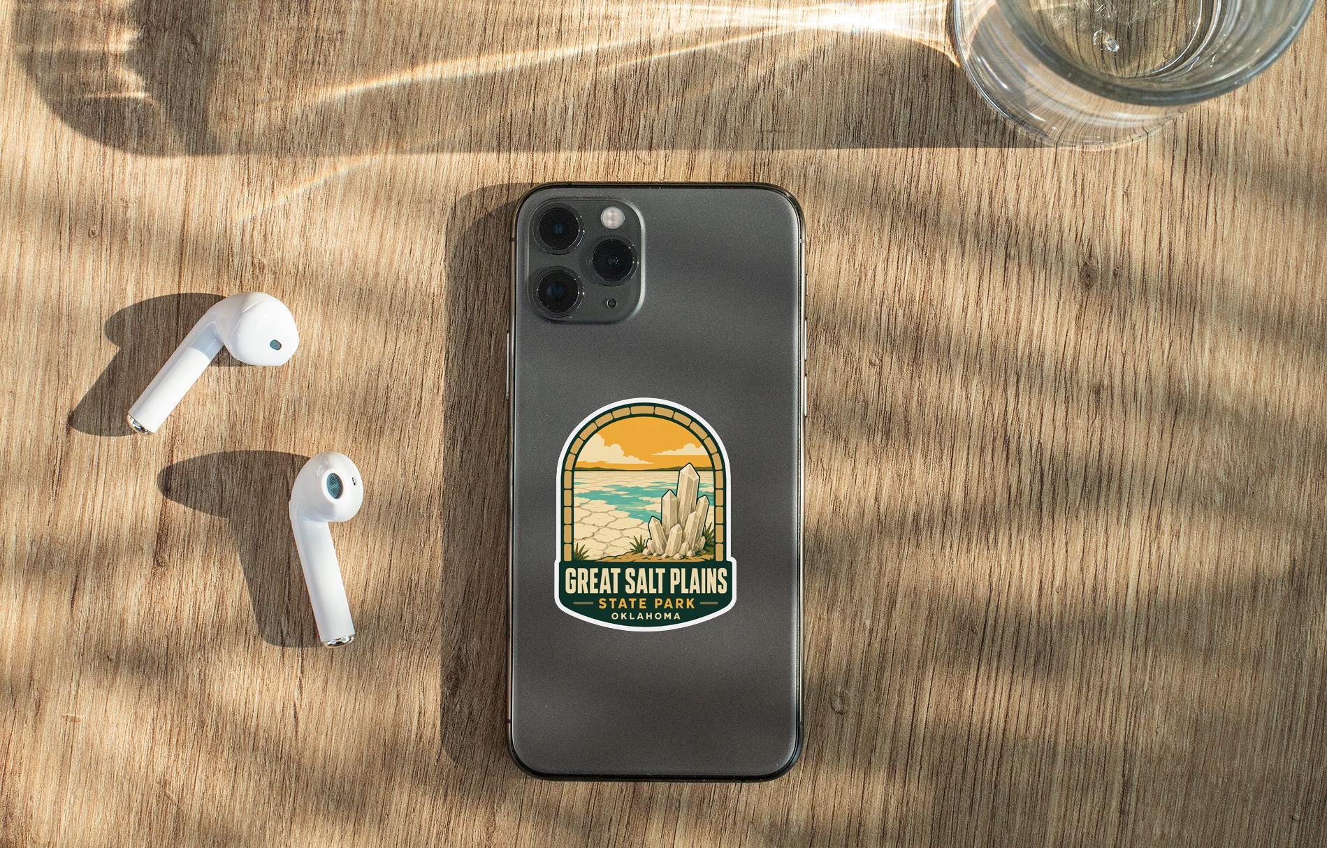 a smartphone with a sticker on the back, placed on a wooden surface alongside a pair of white earbuds and a glass of water.