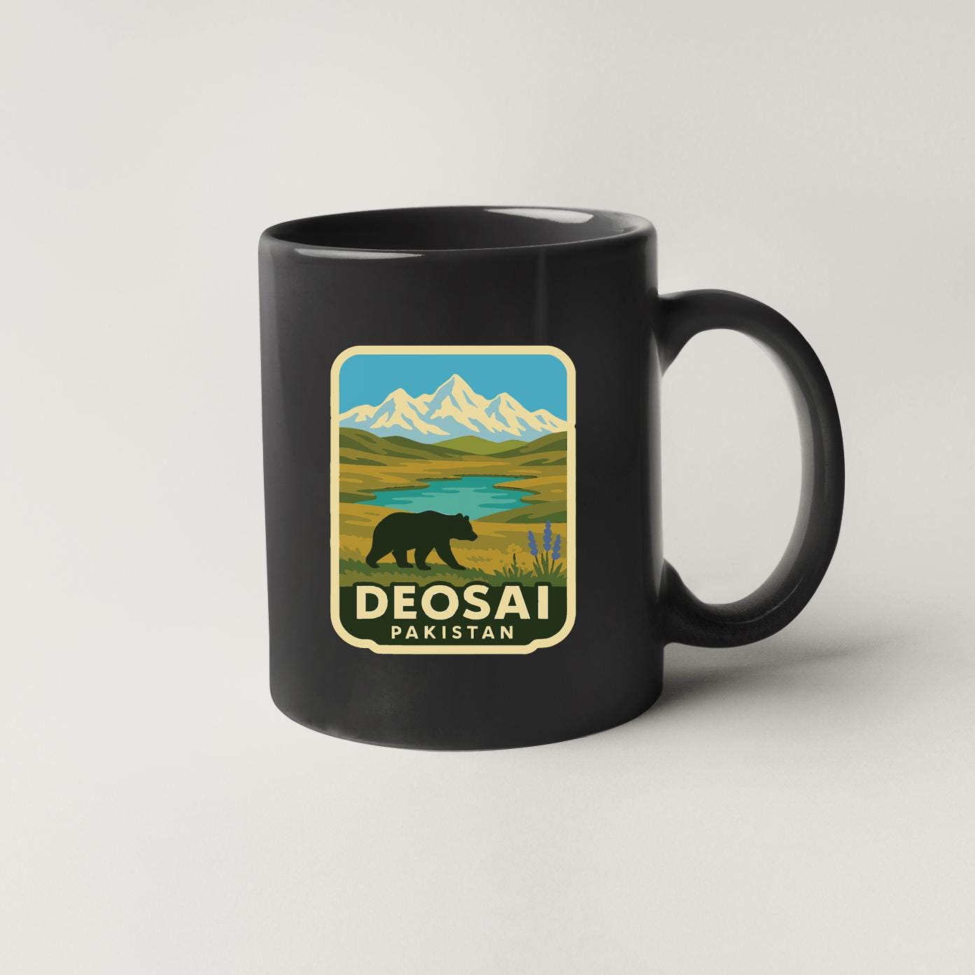 A black coffee mug with a sticker depicting a bear in a mountainous landscape and the text "DEOSAI PAKISTAN" printed on it.