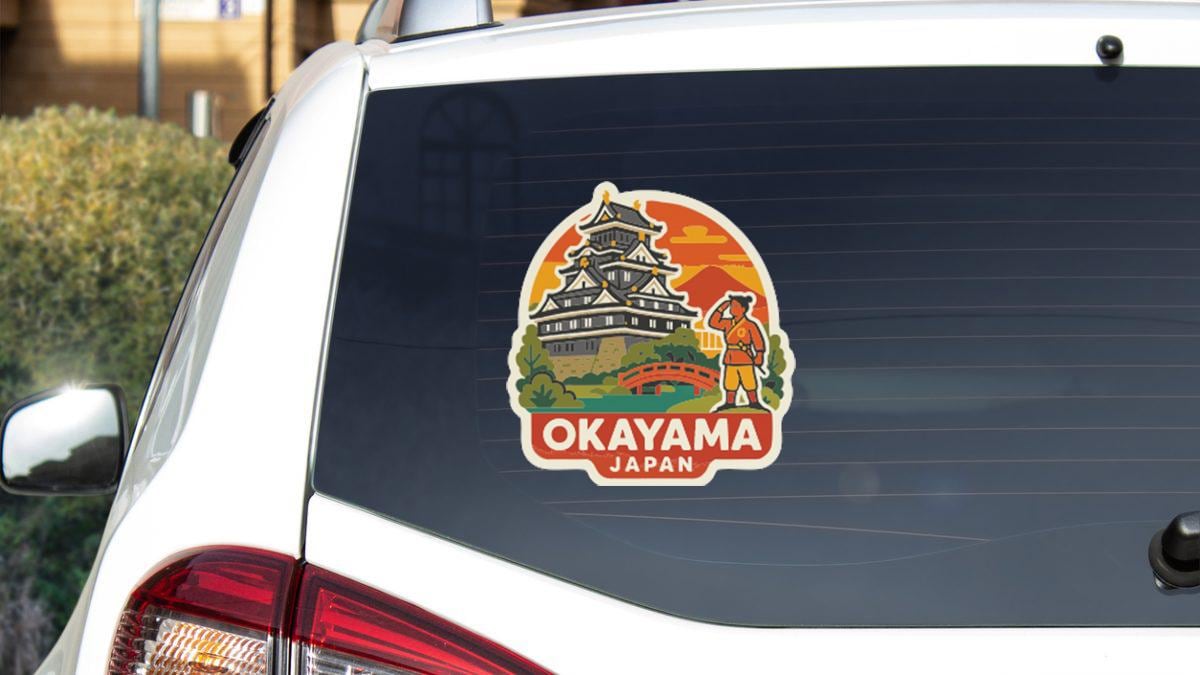 A sticker on the rear window of a car that features an illustration of a castle and a person, along with the text "OKAYAMA JAPAN".