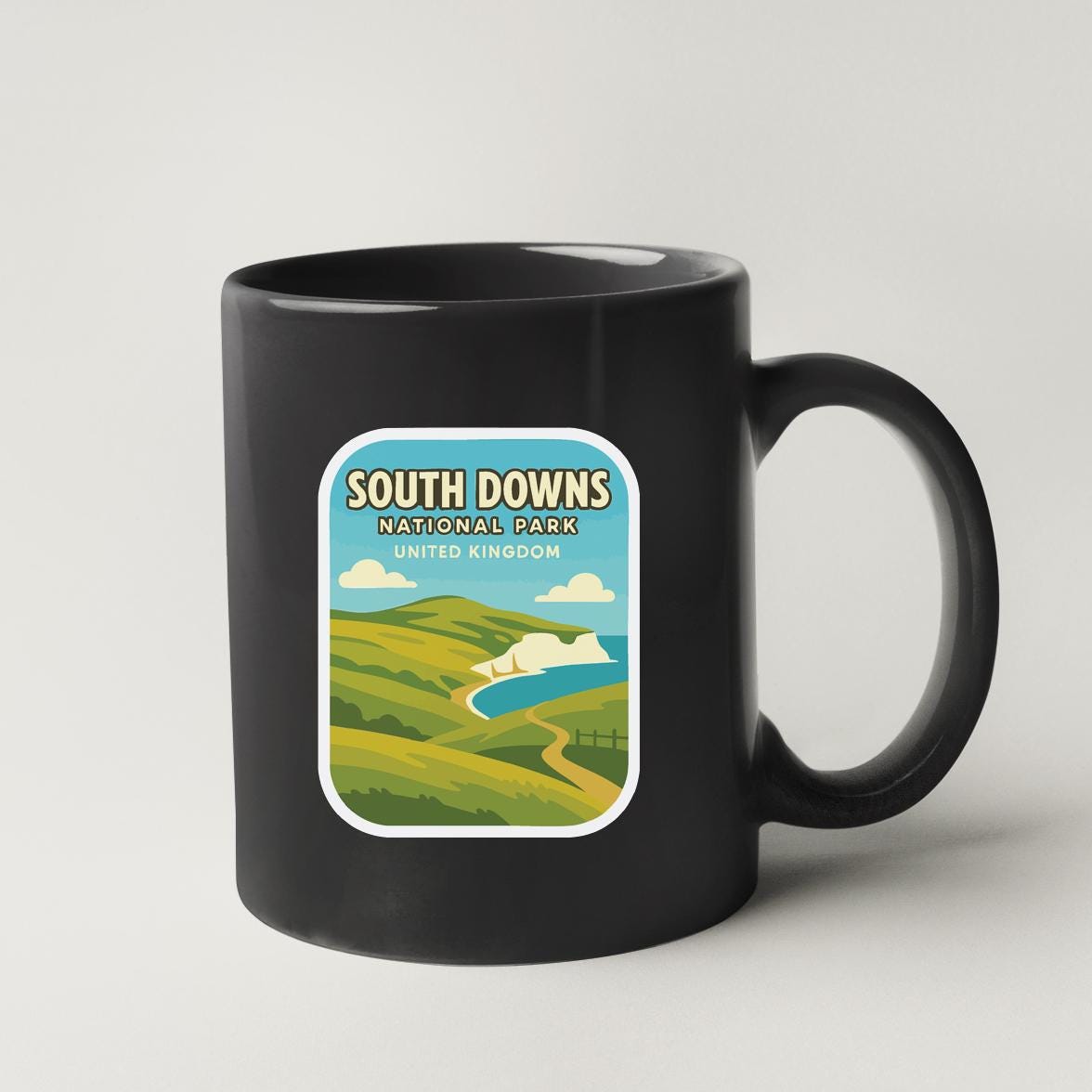 A black coffee mug with a sticker depicting a scenic landscape, including hills, a river, and a castle, along with the text "SOUTH DOWNS NATIONAL PARK UNITED KINGDOM".
