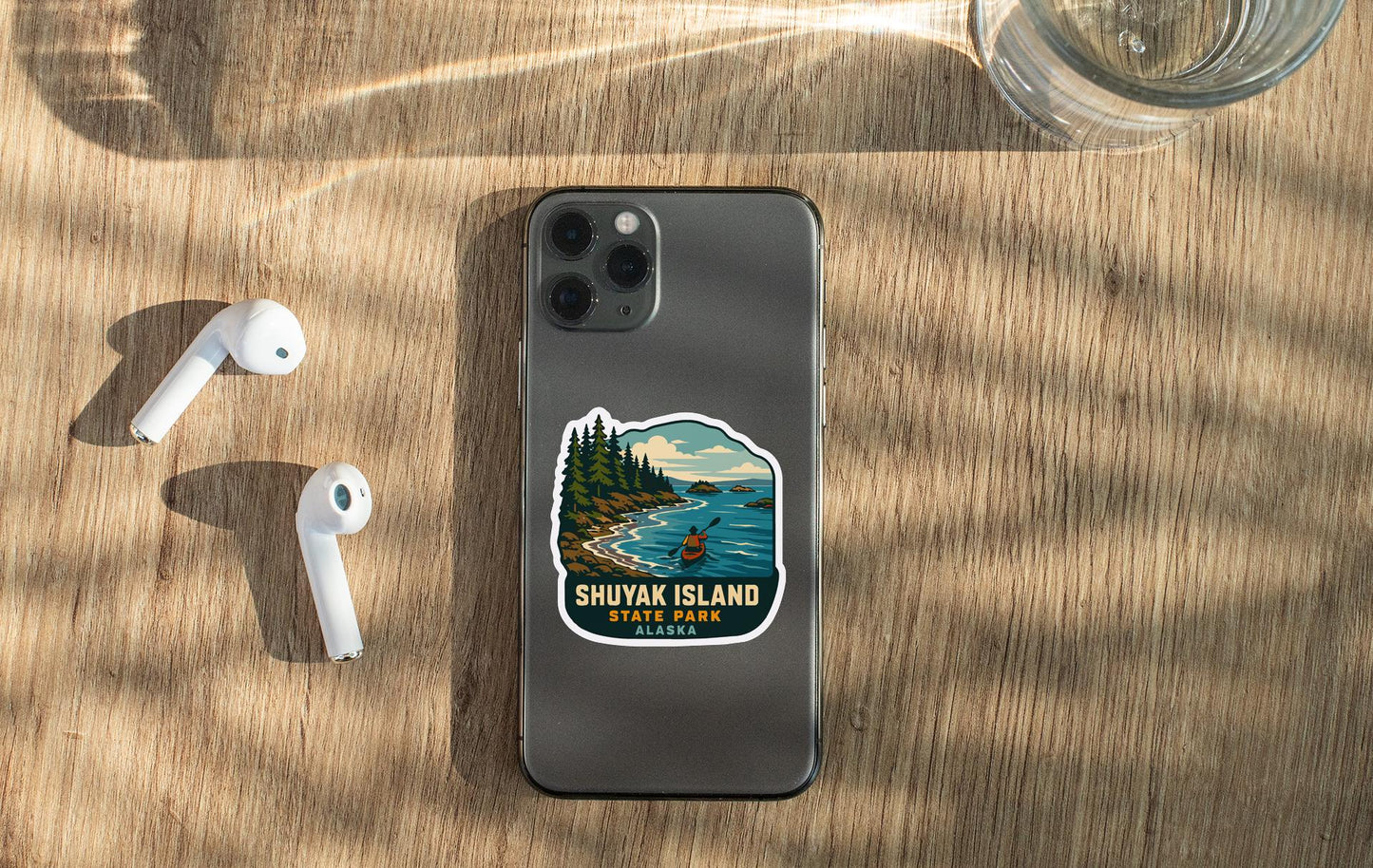 a smartphone with a sticker of Shuvak Island State Park on it, along with a pair of white earbuds and a glass of water on a wooden surface.