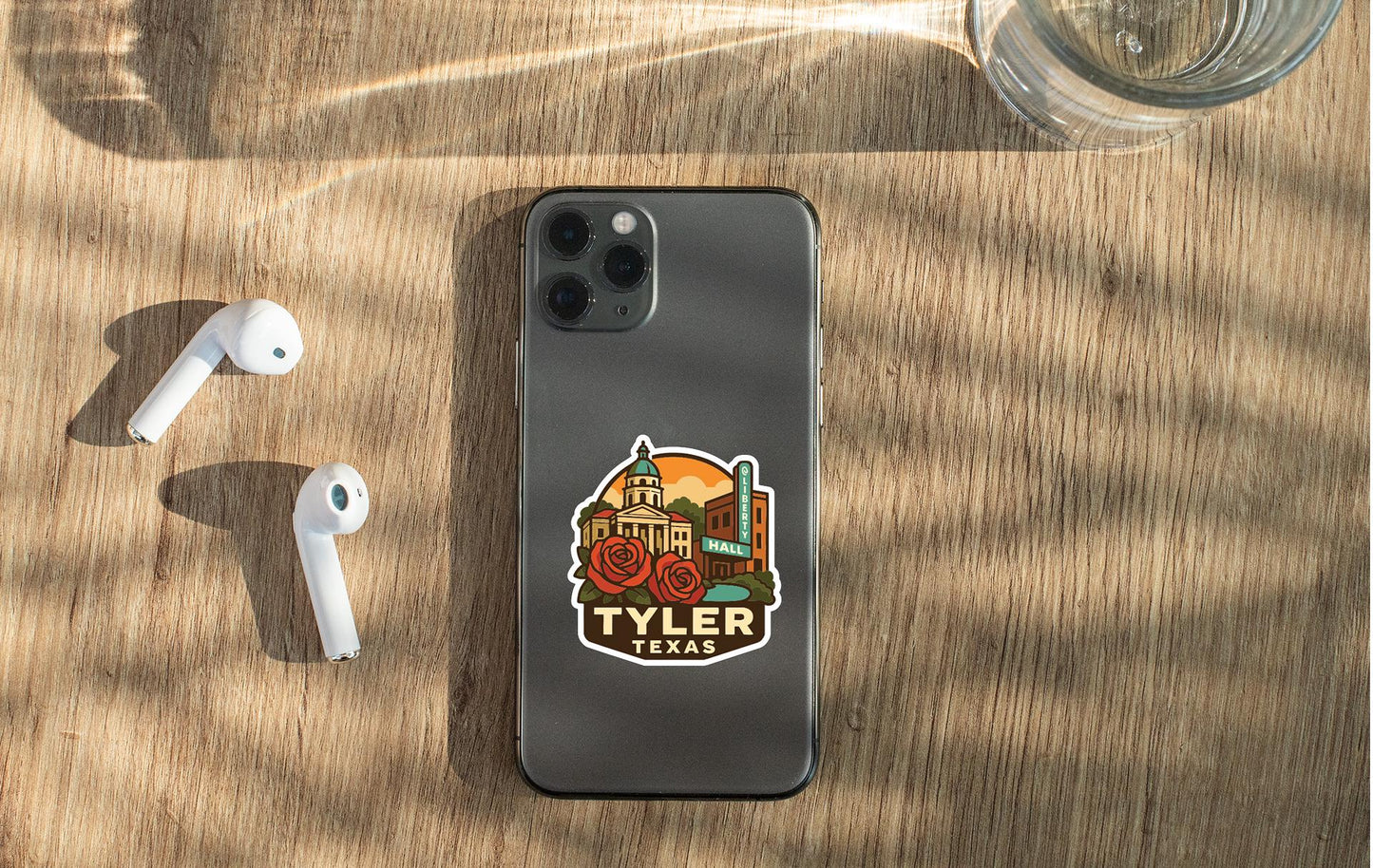a smartphone with a sticker of a landmark, specifically the Tyler, Texas building, placed on its back. The phone is accompanied by a pair of white earbuds and a glass of water, all resting on a wooden surface.