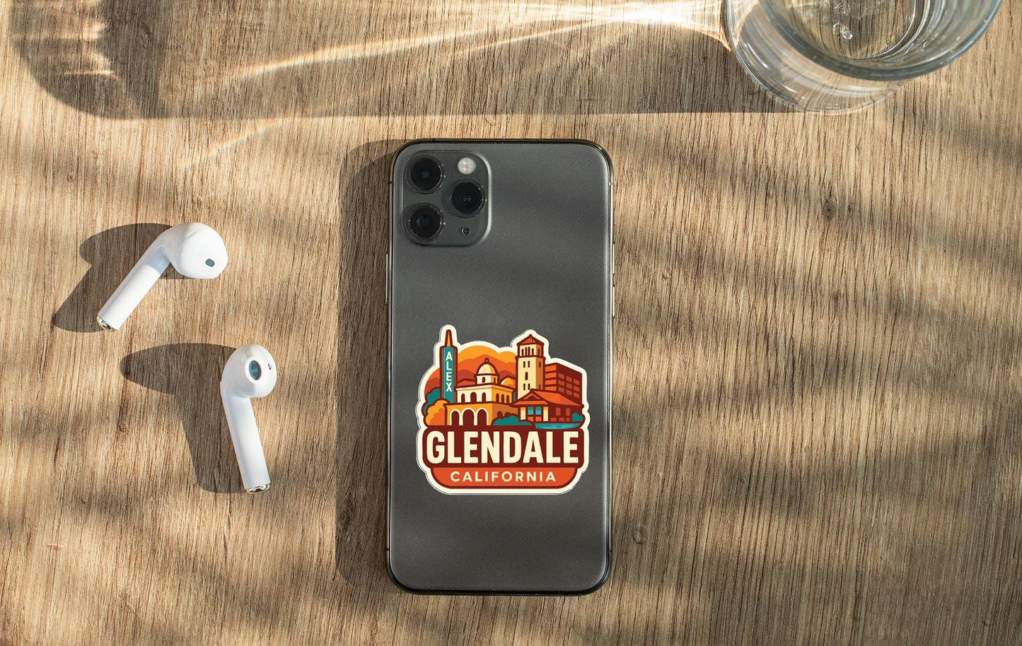 a smartphone with a sticker of Glendale, California, placed on a wooden surface alongside a pair of white earbuds and a glass of water.