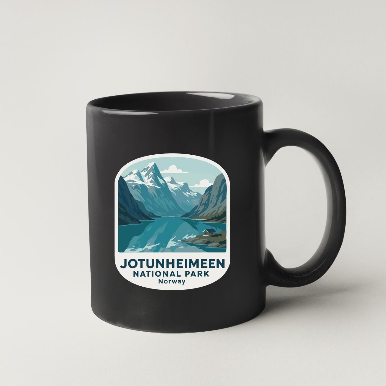 A black coffee mug with a graphic design depicting a scenic mountain landscape and a lake.