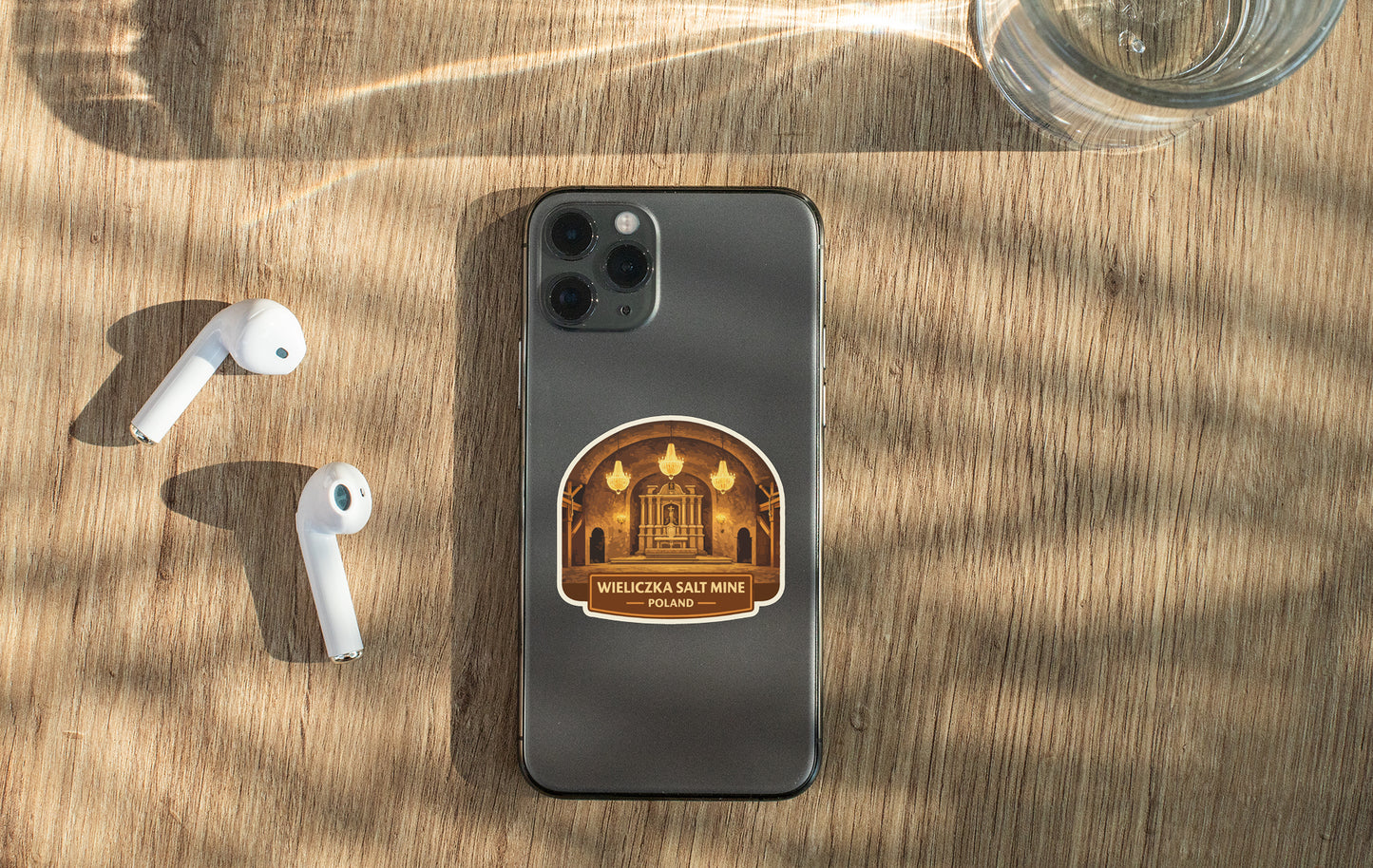 a smartphone with a sticker on the back, placed on a wooden surface alongside a pair of white earbuds and a glass of water.