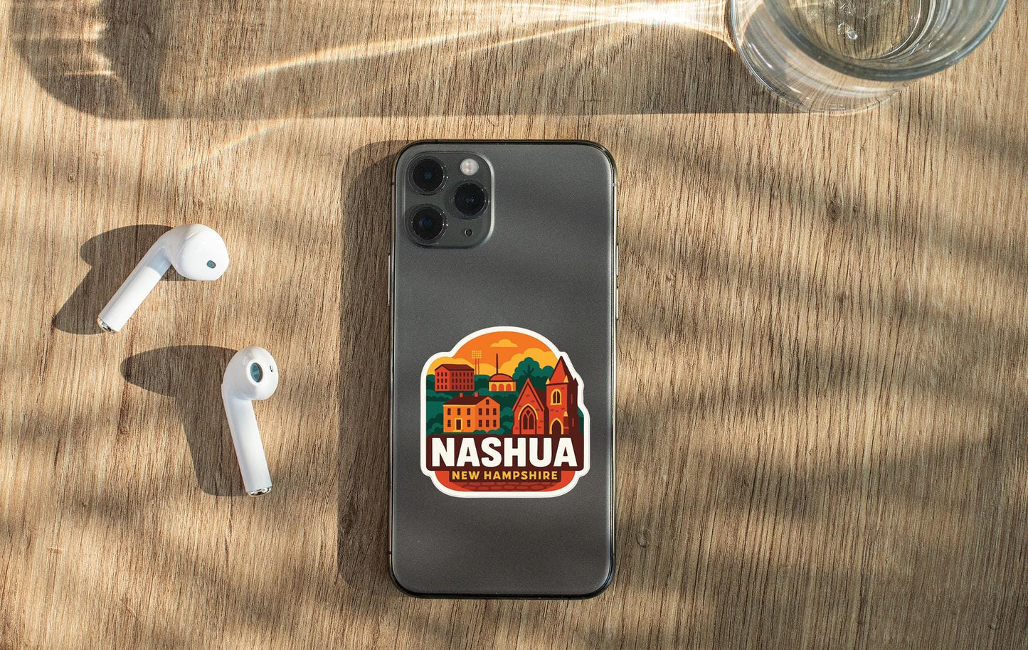 a smartphone with a sticker of Nashua, New Hampshire, on its back, along with a pair of white earbuds and a glass of water on a wooden surface.