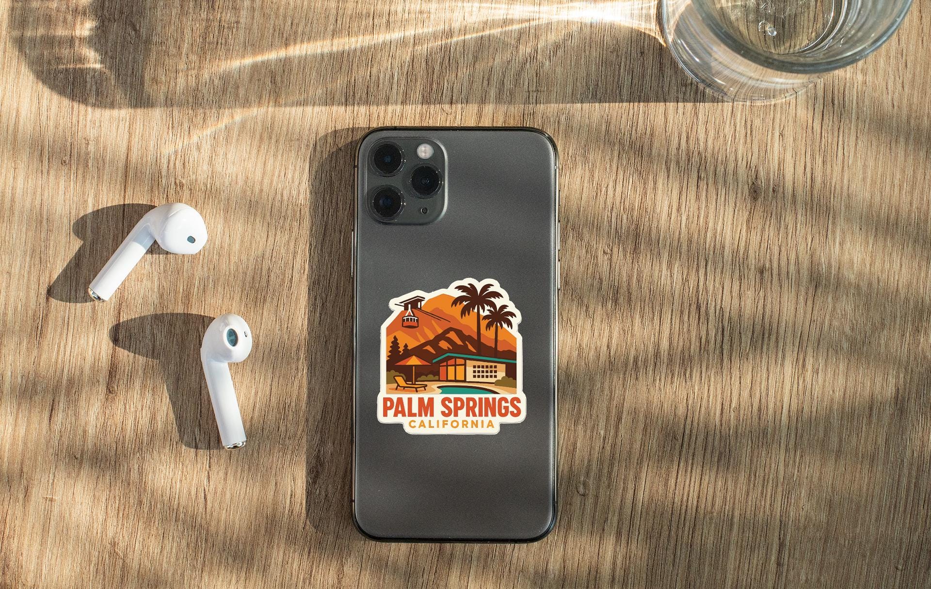a smartphone with a sticker of Palm Springs, California, placed on a wooden surface alongside a pair of white earbuds and a glass of water.